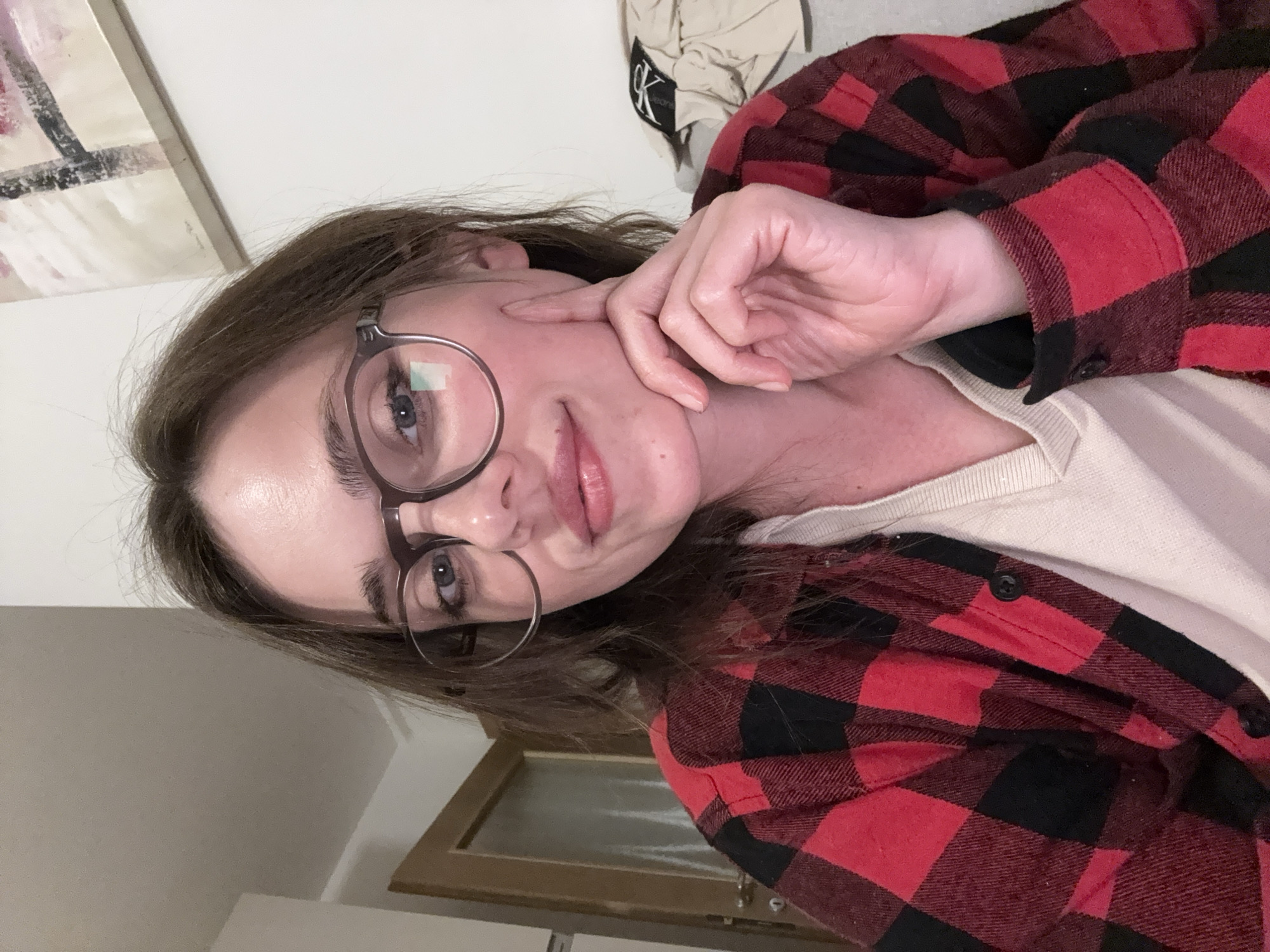 A person with brown hair and glasses is wearing a red and black flannel shirt over a white undershirt. The person is touching their face with their right hand. In the background, there is a painting on the wall and a Calvin Klein jacket. The location is unknown.