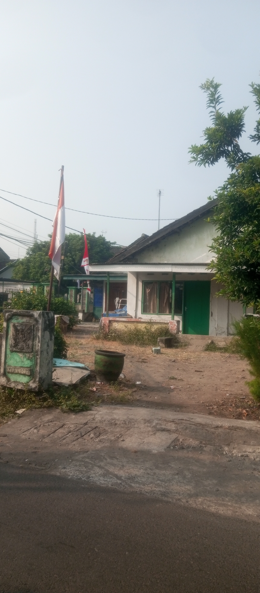 The image shows a building with a green door and windows, with a pitched roof. A flag, which appears to be the Indonesian flag, is flying from a pole in front of the building. There is a concrete structure with some markings to the left of the flag. A large green pot is situated on the ground near the building. The area in front of the building is unpaved and appears to be somewhat dry and dusty, with some sparse vegetation. Trees are visible behind the building and to the right. Power lines are present in the background. The sky is overcast. The scene is likely a residential or public building in Kediri City, Indonesia.