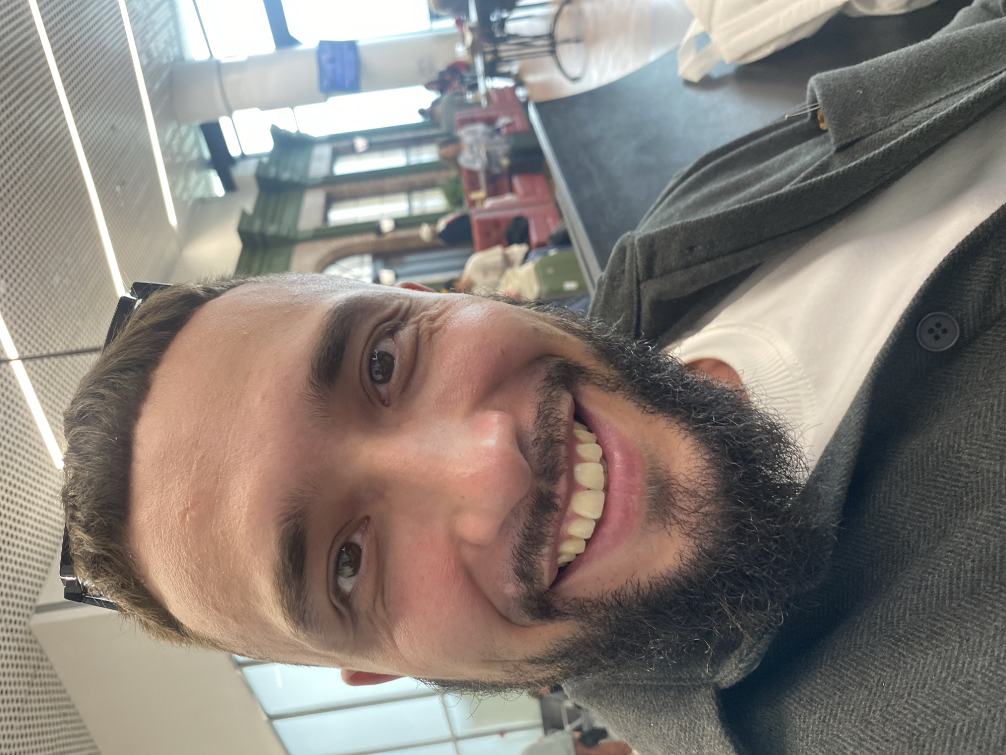 This is a close-up selfie of a man with short brown hair, a beard, and brown eyes, taken indoors. He is smiling widely, showing his teeth, and wearing a grey collared jacket over a white t-shirt. Sunglasses are perched on top of his head. The background appears to be an airport or a large waiting area, with a tiled ceiling and large windows. In the blurred background, there are other people sitting at tables and on benches, and architectural elements that suggest a public transportation hub. The lighting is bright and diffused, likely from overhead fixtures and natural light. There is no visible indication of the specific location being Новое Мартьяново, Russia.