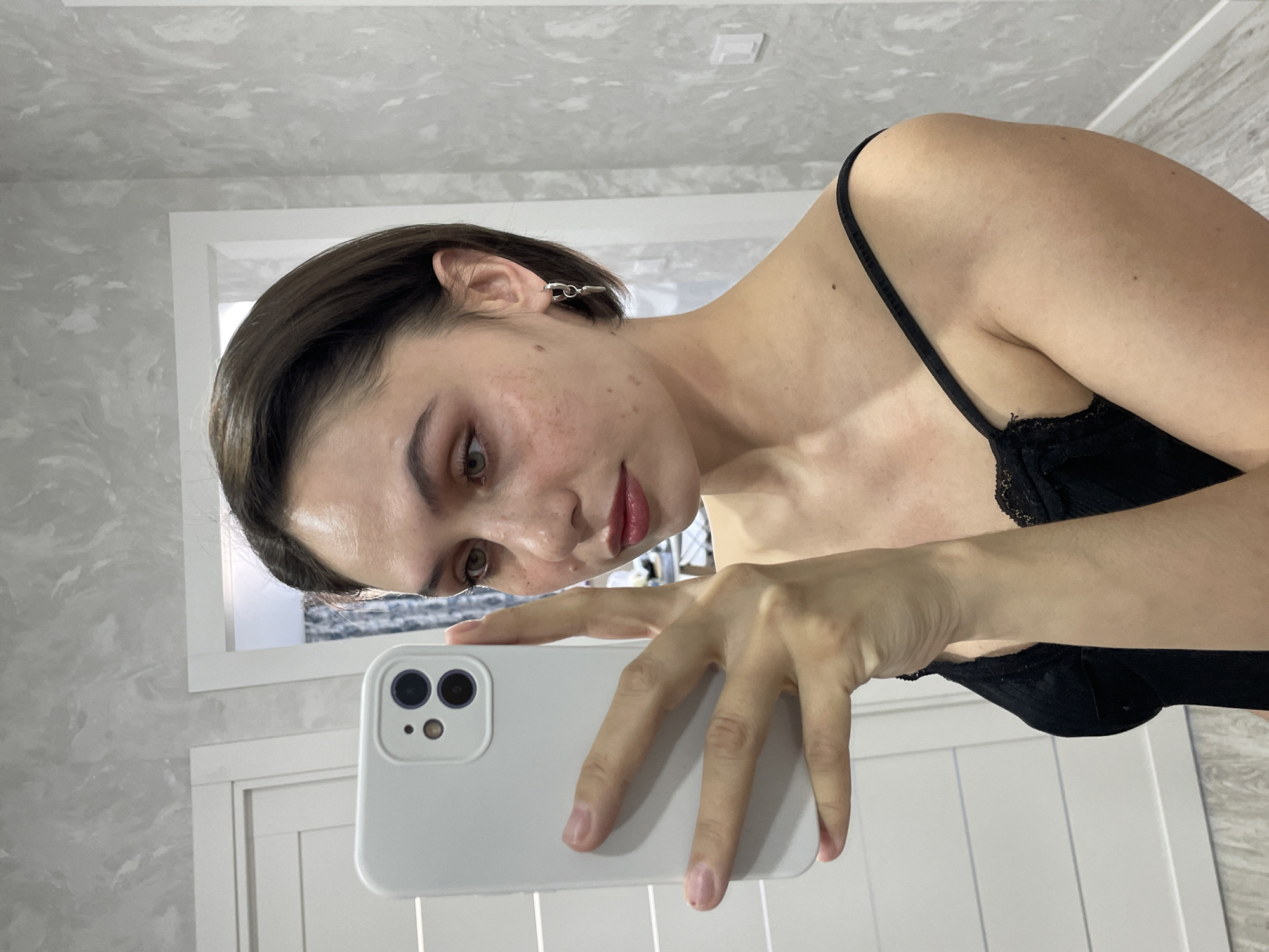 A young woman is the primary subject, captured in a mirror selfie within an indoor setting, likely in Izhevsk, Russia. She has short, dark brown hair, light-colored eyes, and visible freckles or blemishes on her face. She wears a black, lace-trimmed bralette or camisole, and a distinct silver-toned earring is visible in her left ear.
The woman is actively taking the selfie, holding a white smartphone – likely an iPhone model with a dual-lens camera – in her right hand. Her head is tilted slightly, and her gaze is directed towards her reflection in the phone camera, presenting a soft, neutral expression with a subtle pout.
The background elements include a light-colored wall featuring a mottled grey and white pattern, possibly textured paint or wallpaper, adding to the contemporary feel of the room. Behind her, a white-framed mirror is visible, reflecting parts of the room, including the upper section of a white panel door or cabinet situated beneath the mirror. The lighting in the scene appears to be soft and ambient, suggesting an indoor environment without direct sunlight, making the specific time of day indistinguishable. There is no visible text in the image.