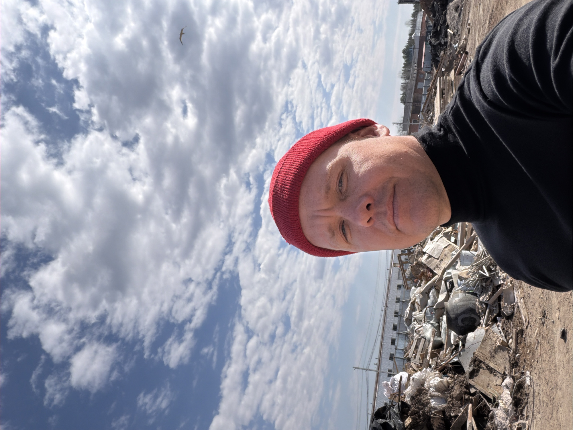 A man is captured in a selfie taken outdoors during the daytime. He is positioned in the foreground, wearing a red beanie and a black long-sleeved shirt or turtleneck, looking directly at the camera with a neutral expression.
The sky above him is partly cloudy with patches of blue. A single bird is visible flying in the upper left portion of the frame. In the background, to the man's right, there is a large, messy pile of diverse debris, including wood, metal, plastic, and other discarded materials, suggesting a junkyard, construction site, or industrial waste area. The ground beneath this pile and in the immediate foreground appears to be dirt.
Further in the distance, behind the debris pile, a long, low-rise building with a corrugated metal roof and multiple windows can be seen. Beyond this structure, a line of trees extends horizontally. The scene depicts an industrial or semi-rural outdoor setting, consistent with the provided location context of Chaikovsky, Russia. No visible text is present in the image.