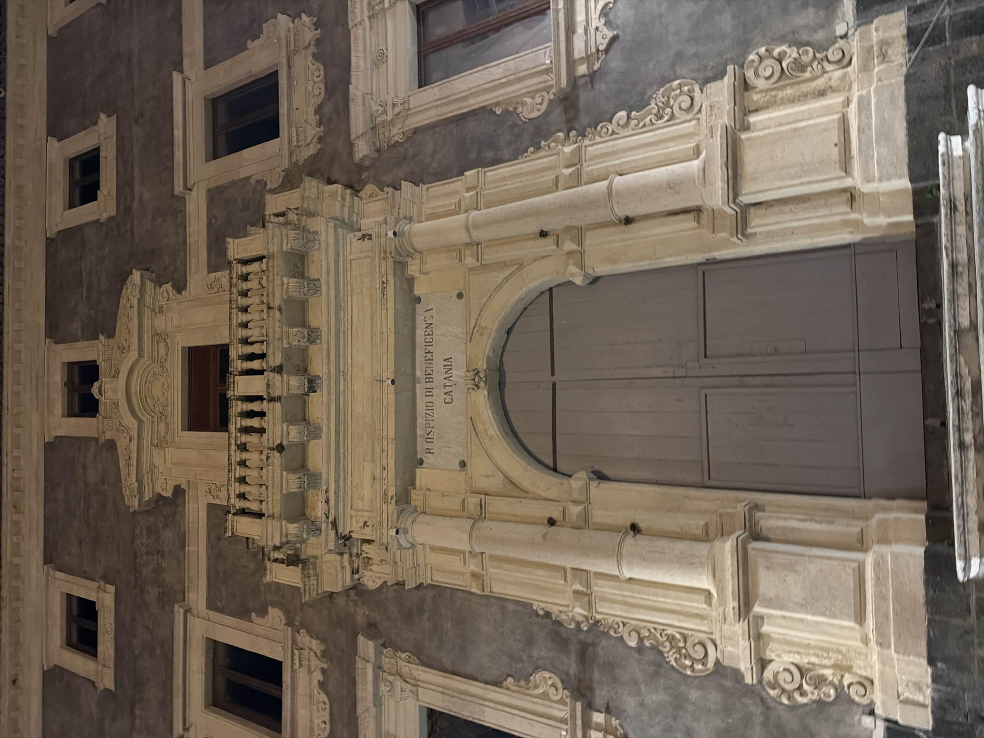 The image depicts the façade of the R. Ospizio di Beneficienza in Catania, Italy. The building is characterized by its ornate Baroque architectural style, featuring elaborately decorated windows and an arched entrance. The façade is predominantly gray with light-colored stone or stucco details, including decorative moldings and sculptural elements. The architectural style is consistent with historical buildings found in Catania.