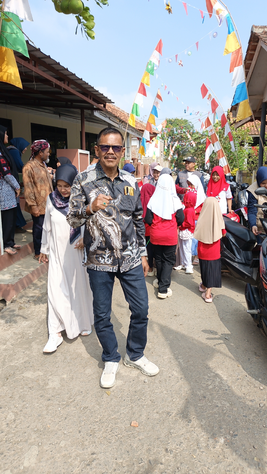The image depicts an outdoor scene in Linggasirna, Indonesia, likely a village street, adorned with numerous colorful flags and pennants strung across the pathway, suggesting a festive or celebratory occasion. The foreground features a man in his 50s, wearing sunglasses and a patterned black shirt, blue jeans, and white sneakers, posing with his left hand on his hip. He is the central subject and appears to be looking towards the camera. To his left, a woman in a long, light-colored dress and headscarf is walking away from the camera. Behind them and to the right, a group of people, including several children in red and white attire, are gathered. Some individuals are wearing hijabs. Various motorcycles are parked along the side of the street, and buildings with tiled roofs are visible in the background. The ground is paved with rough asphalt or gravel. The sky is clear and blue. The overall impression is one of a community gathering on a bright, sunny day.