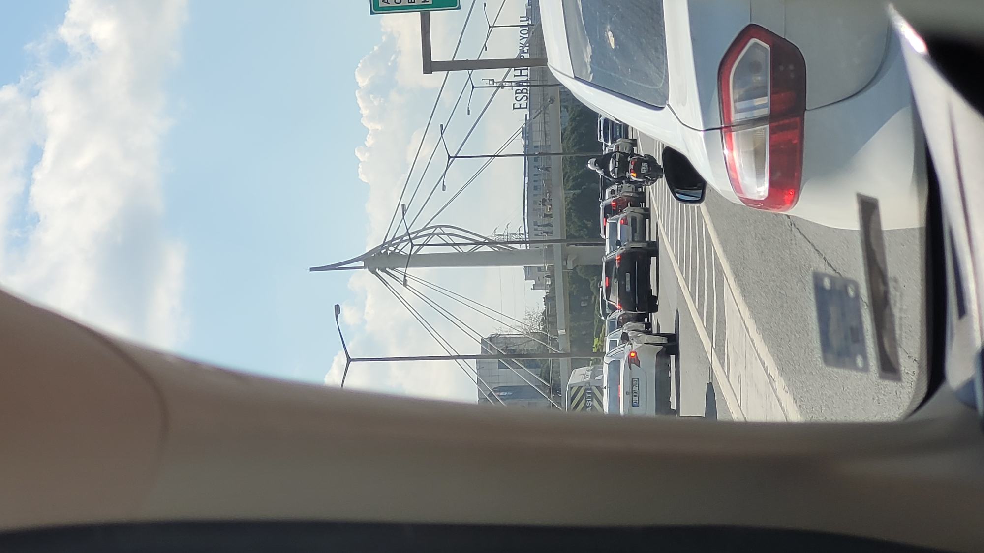 The image, likely taken from inside a vehicle, shows heavy traffic on a road in Küçükçekmece, Turkey. Several cars are visible, lined up in multiple lanes. A white car's taillight is prominent in the foreground. In the background, a distinctive bridge structure with a central tower supported by cables is apparent. Roadside infrastructure, including streetlights and signage, are also visible. A sign appears to show "ESBAH PEKYOLU". The sky is bright with scattered clouds.