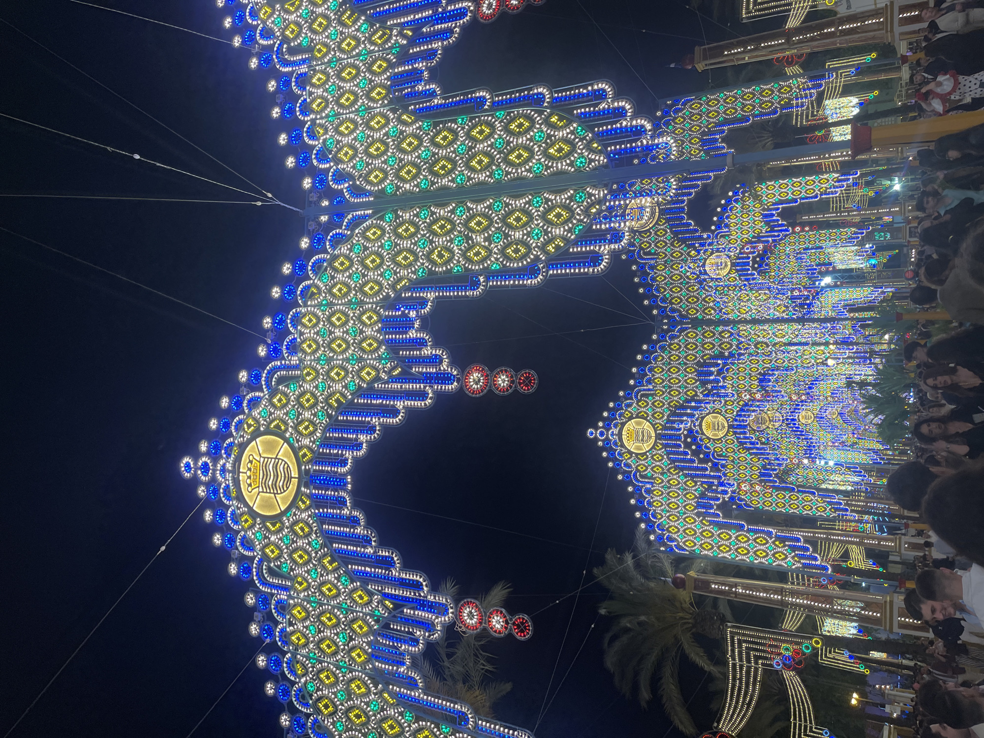 This nighttime photograph shows a street in Jerez De La Frontera, Spain, decorated for a festival. Illuminated archways stretch across the street, composed of numerous small light bulbs arranged in geometric patterns, primarily diamonds, with colors of blue, yellow, white, and green. The archways feature central decorative medallions. At the edges of the archways, lines of blue bulbs form the outlines. Some red circular decorations are visible as well. Crowds of people line the sides of the street, and palm trees are interspersed between the arches. The scene is dark, illuminated primarily by the festive lighting.