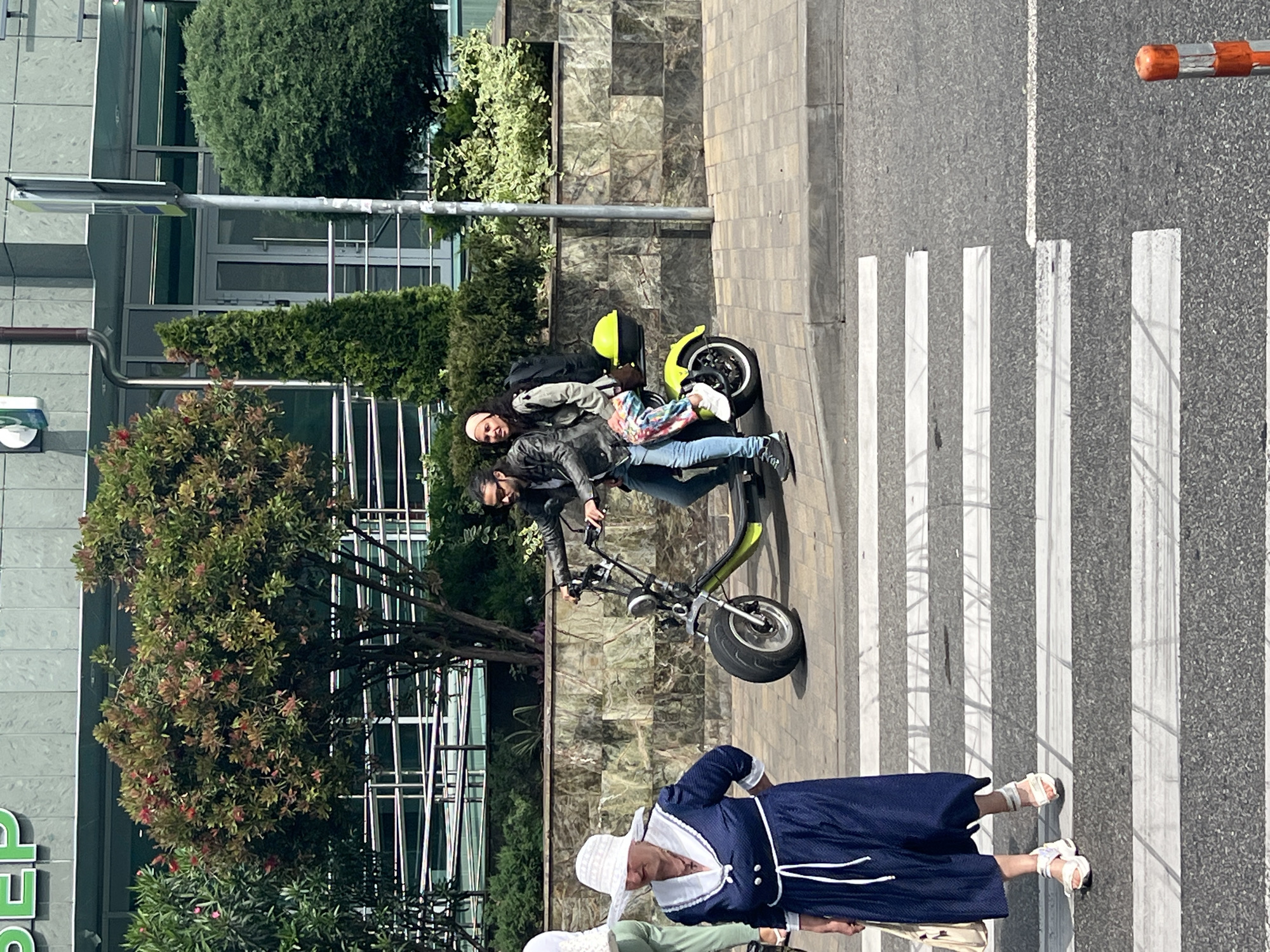 In Sochi, Russia, on a sunny day, an older woman in a navy blue dress and a white hat stands on a pedestrian crossing. A younger couple is riding a lime green scooter. The man is driving, wearing sunglasses and a black leather jacket, while the woman behind him wears a light-colored jacket and colorful pants. They are on the sidewalk adjacent to the pedestrian crossing. The background shows a building with glass windows and some greenery, including trees and bushes.