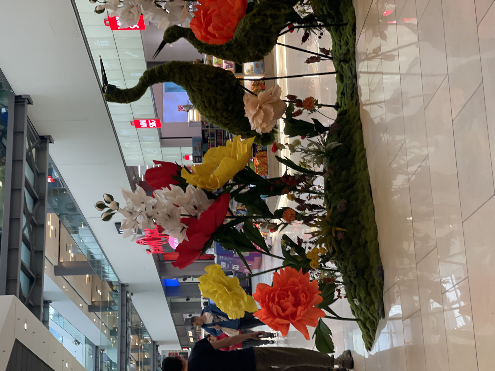 The image shows a mall interior featuring an elaborate floral arrangement. The arrangement includes large artificial flowers in red, yellow, orange, and white, along with moss-covered bird-like figures. People are walking in the mall, visible in the lower part of the frame. The mall has multiple levels with shops and glass railings. It is illuminated with artificial lighting. The location is inside a mall in Sofia, Bulgaria, based on signage visible in the image.