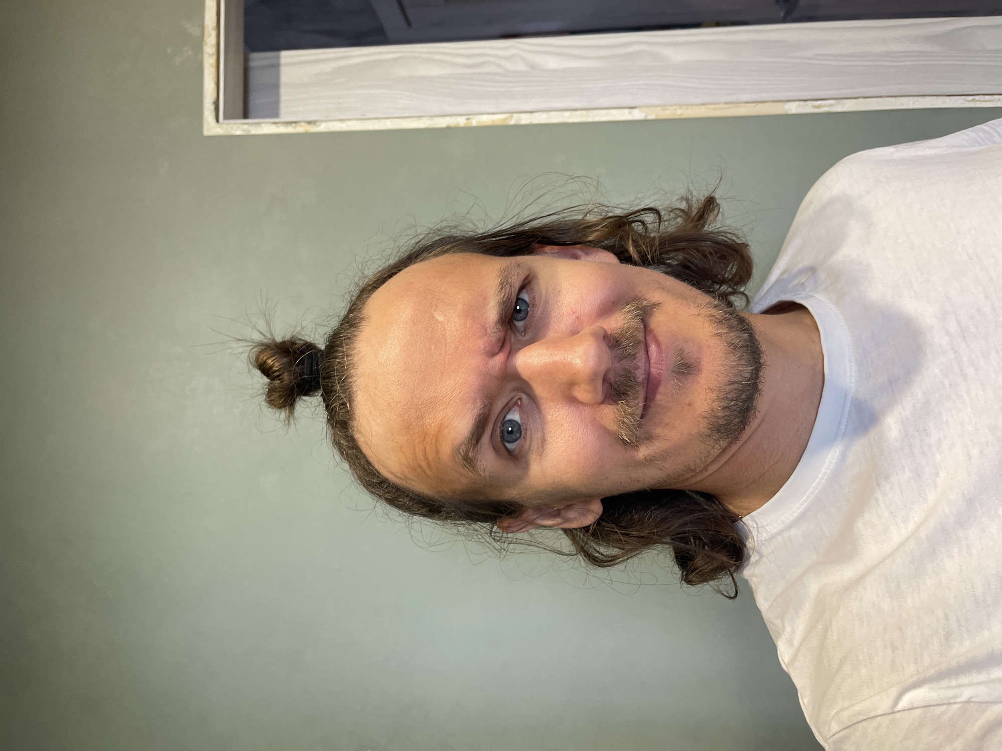 A close-up portrait, correctly oriented, features a man looking directly at the camera. He has long, wavy dark brown hair pulled back into a small bun at the top of his head. He has light blue eyes and a light brown mustache and beard stubble. His expression shows a slight, almost mischievous smirk, with his eyebrows subtly raised and faint horizontal lines visible on his forehead. He is wearing a plain white crew-neck t-shirt. The background consists of a solid, light greenish-gray wall. In the upper right corner, a small section of a light wooden frame is visible, bordering an indistinct darker area, possibly a cabinet or mirror. The lighting is soft and even, indicating an indoor setting. No text is visible in the image.