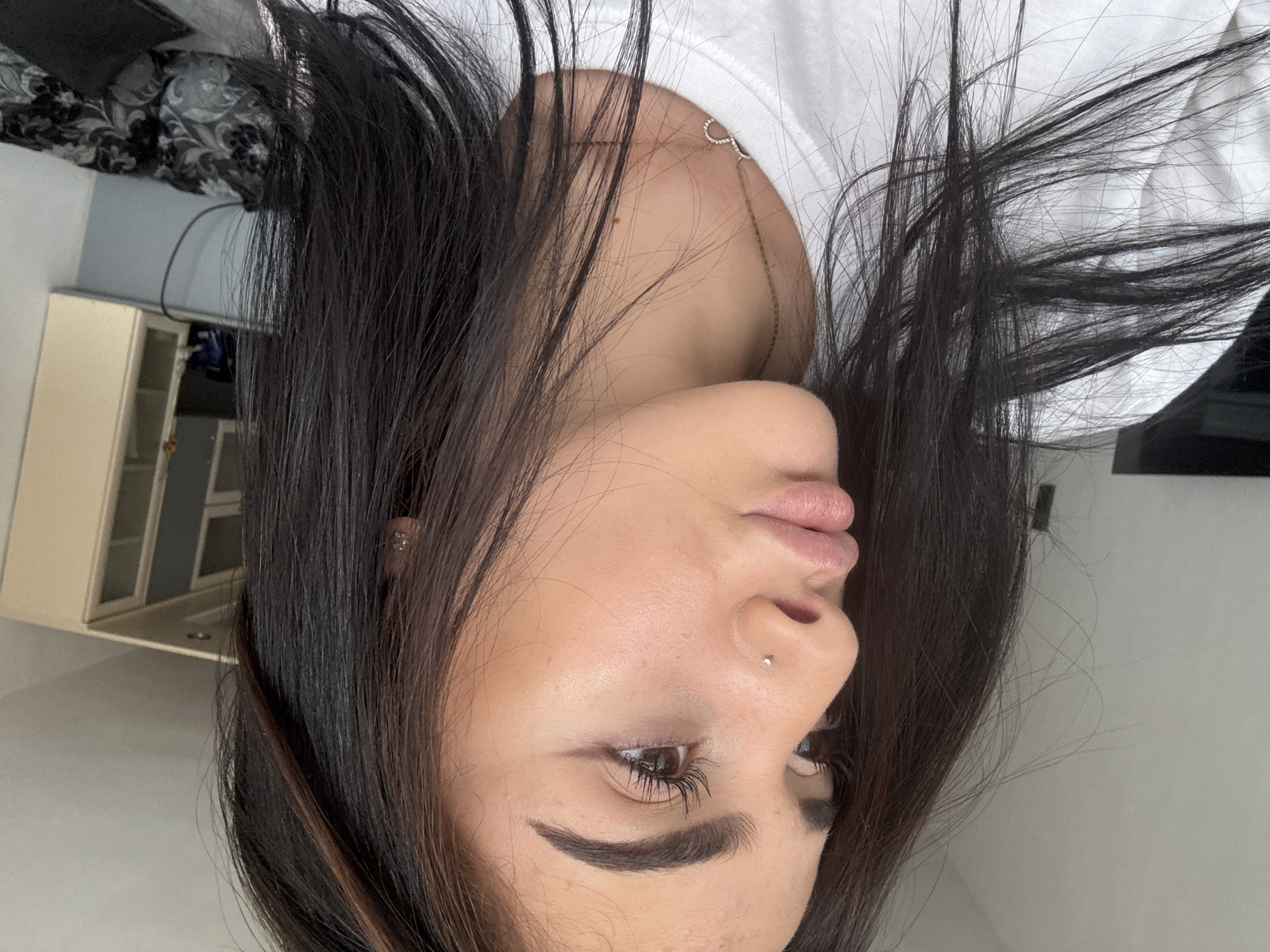 A close-up portrait of a young woman with long, dark hair partially obscuring her face. Her skin tone is fair, and she has prominent eyebrows and full lips. She is wearing a white top, and a necklace is visible. A small stud earring is present in her ear, and a piercing adorns her nose. The background includes a cabinet and a patterned surface, suggesting an indoor setting. The location is in the city of Unknown, Unknown.