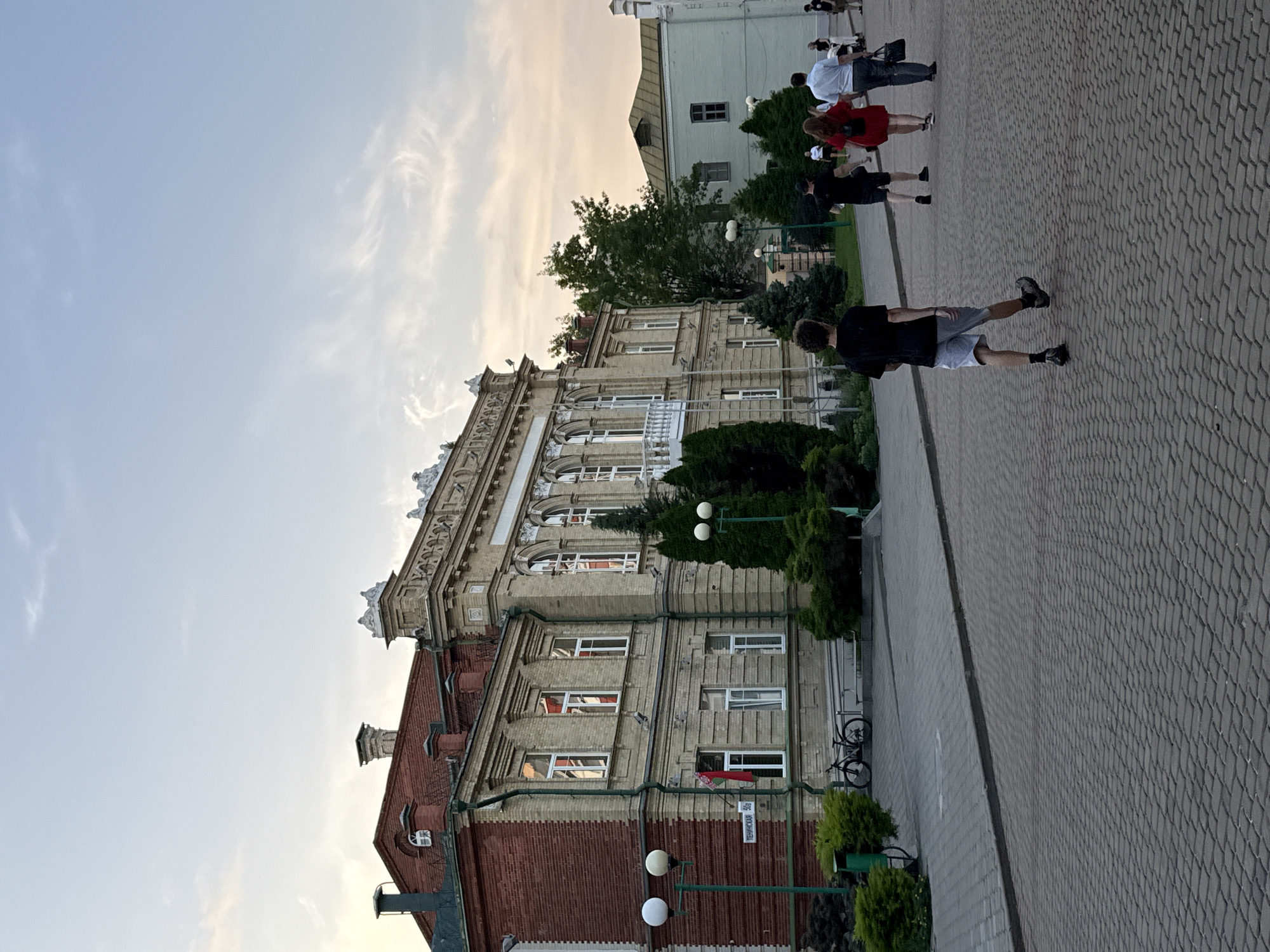 The image depicts a street scene in Mahilyow, Belarus, with a focus on a large, ornate yellow building constructed of brick and stone. The building features arched windows, decorative cornices, and a white balcony. To the left of the yellow building is a smaller red brick building with a green roof. A flag, appearing to be the flag of Belarus, hangs from the yellow building. A bicycle is parked on the sidewalk in front of the building.
Several people are walking on the cobblestone sidewalk in the foreground, moving away from the camera. One individual in the immediate foreground wears a black t-shirt and grey shorts. Further ahead, a group includes a woman in a red dress, a man in a white shirt and dark pants, and another man in a black t-shirt and dark shorts. The background shows trees and other buildings, with a cloudy sky overhead. The overall impression is of a late afternoon or early evening in an urban setting.