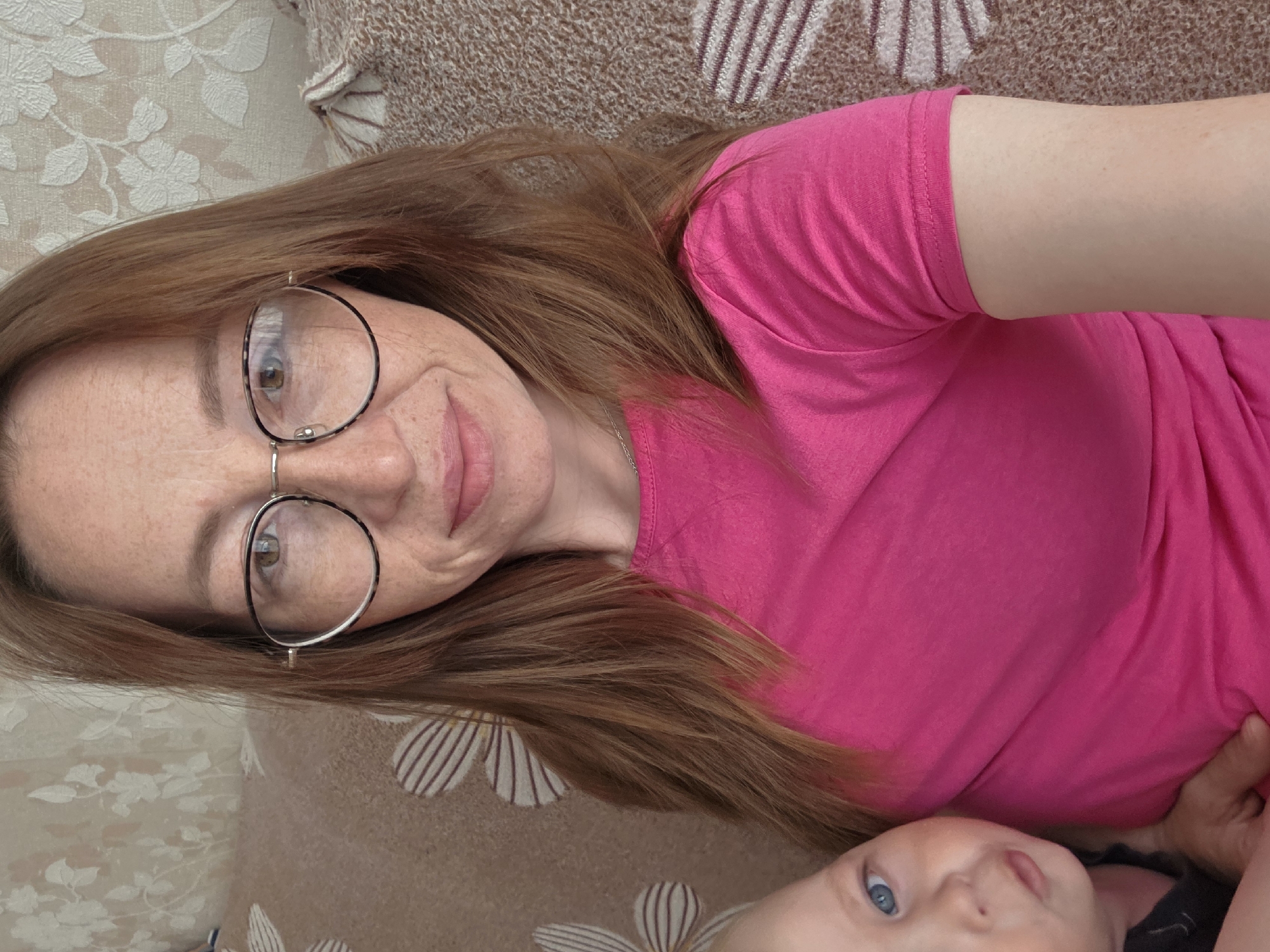 A woman with red hair and glasses, wearing a pink shirt, is lying down with a baby. The scene is indoors, on a patterned couch or bed.