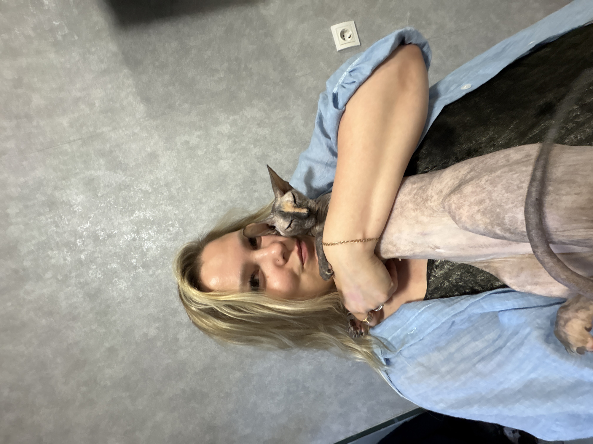 A woman with blonde hair is holding a hairless cat. The woman is wearing a blue button-down shirt. The cat is gray and tan. The background is a gray wall with an electrical outlet. The location is possibly in Марковский, Russia.