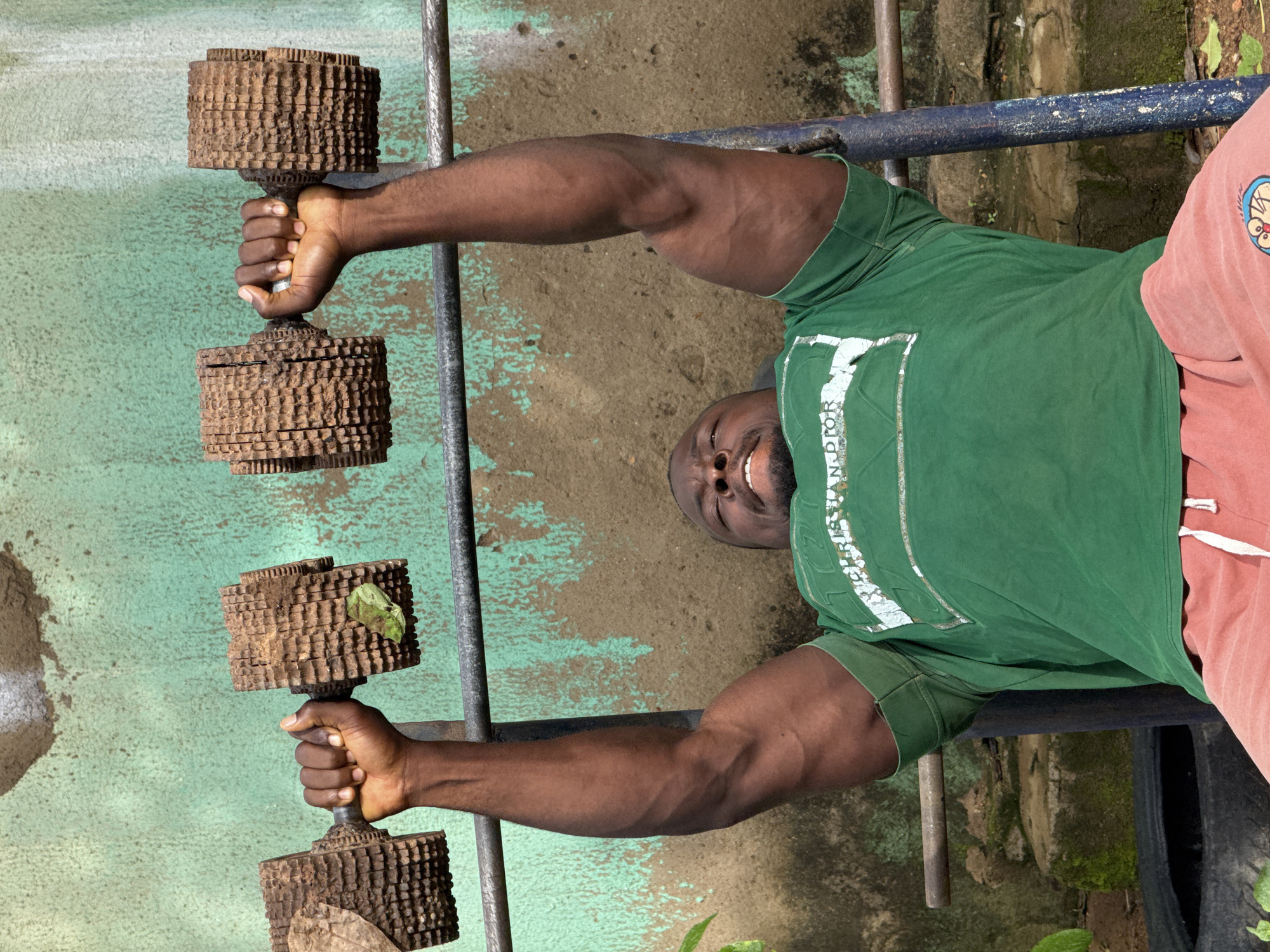 A man is performing bicep curls with a pair of dumbbells. The dumbbells are unusual, made from stacked circular wooden or metal objects, possibly gears or sprockets, giving them a rustic and robust appearance. The man is muscular and wearing a green t-shirt with white lettering and pinkish-orange shorts. He is smiling and appears to be in good spirits. The background suggests an outdoor setting, possibly a gym or workout area, with a textured, painted wall in shades of green and brown. A metal bar runs horizontally behind him. The lighting indicates it is daytime. The scene is set in Akwanga, Nigeria. The visible text on his shirt reads "CHRISTIAN DIOR".