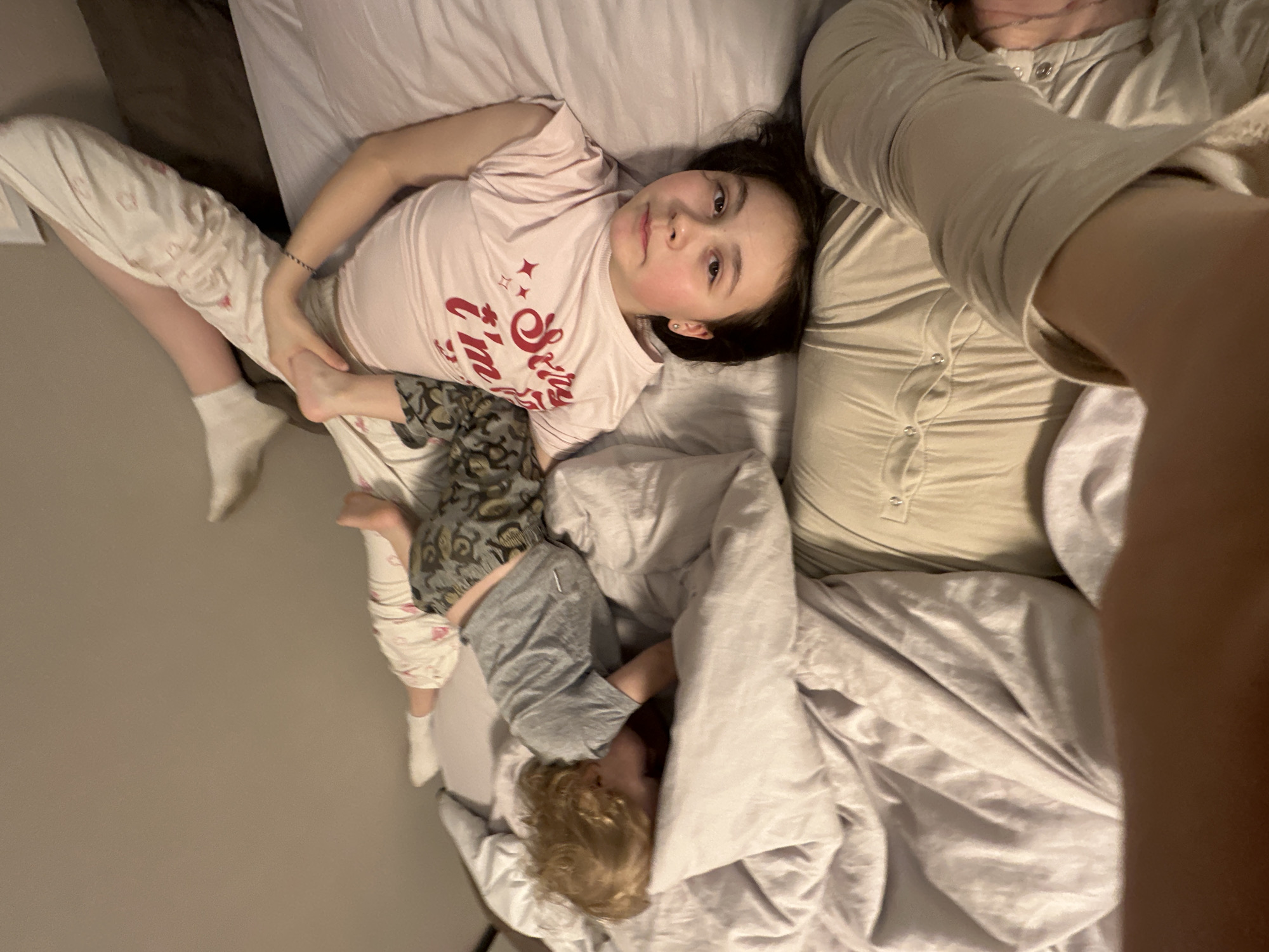 Three people are seen relaxing on a bed indoors in Votkinsk, Russia, captured from a high-angle perspective, likely a selfie taken by an adult. The main subject is an older child, a girl with dark hair, lying on her back, looking directly at the camera with a neutral expression. She wears a light pink t-shirt with red text that reads "Sassy & I'm Cute" and patterned pajama pants. A small black bracelet is visible on her left wrist.
To her lower right, a younger child with blonde, curly hair is partially visible, mostly covered by a light grey duvet, wearing grey shorts. The younger child's face is obscured. An adult is partially visible on the right side of the frame, wearing a light-colored long-sleeved top with button details, with an arm and leg visible, suggesting they are lying down as well and holding the camera. The bed is covered with light-colored sheets and pillows, creating a relaxed, intimate atmosphere. The lighting suggests daytime or early evening.