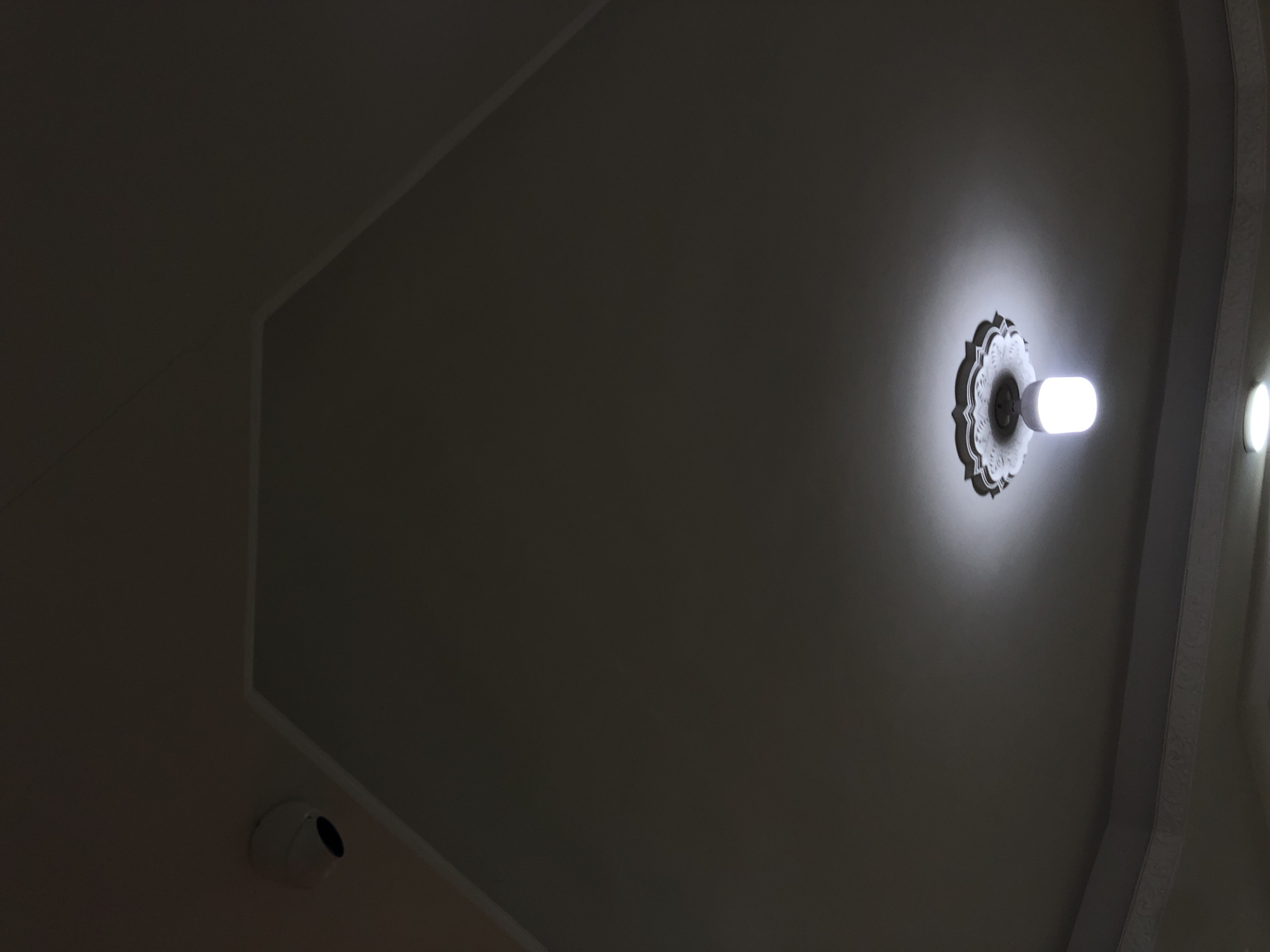 The image shows a dimly lit ceiling. A circular decorative light fixture is mounted near the upper right, emitting a bright light. A security camera is visible in the lower left. The ceiling has a geometrically shaped design feature created by recessed lines. No people are visible. The location in Juan Griego, Venezuela, is not discernible from this image alone.