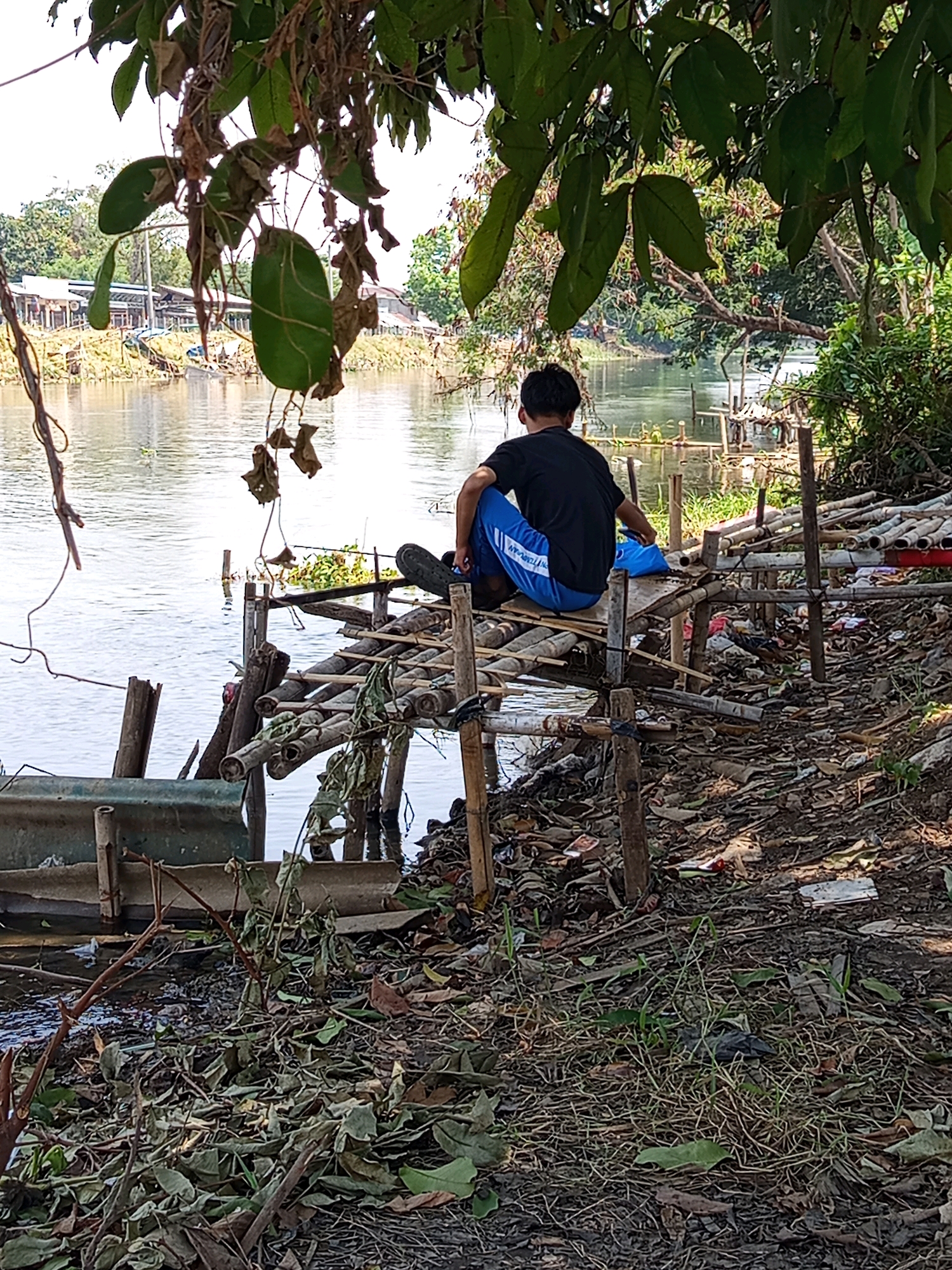 In Karawang, Indonesia, a young male with dark hair, wearing a black t-shirt and blue pants with white stripes, is seated on a makeshift wooden platform by a body of water. He is facing away from the camera, looking towards the river. The platform is constructed from bamboo poles and appears weathered and possibly unstable. Next to him on the platform is a blue plastic bag. The area surrounding the platform is cluttered with debris, including dried leaves, plastic waste, and what appear to be discarded bamboo poles. The river itself is calm and reflects the surrounding trees and sky. In the background, there are some buildings and more vegetation. The overall impression is of an informal settlement or a neglected area along the riverbank.