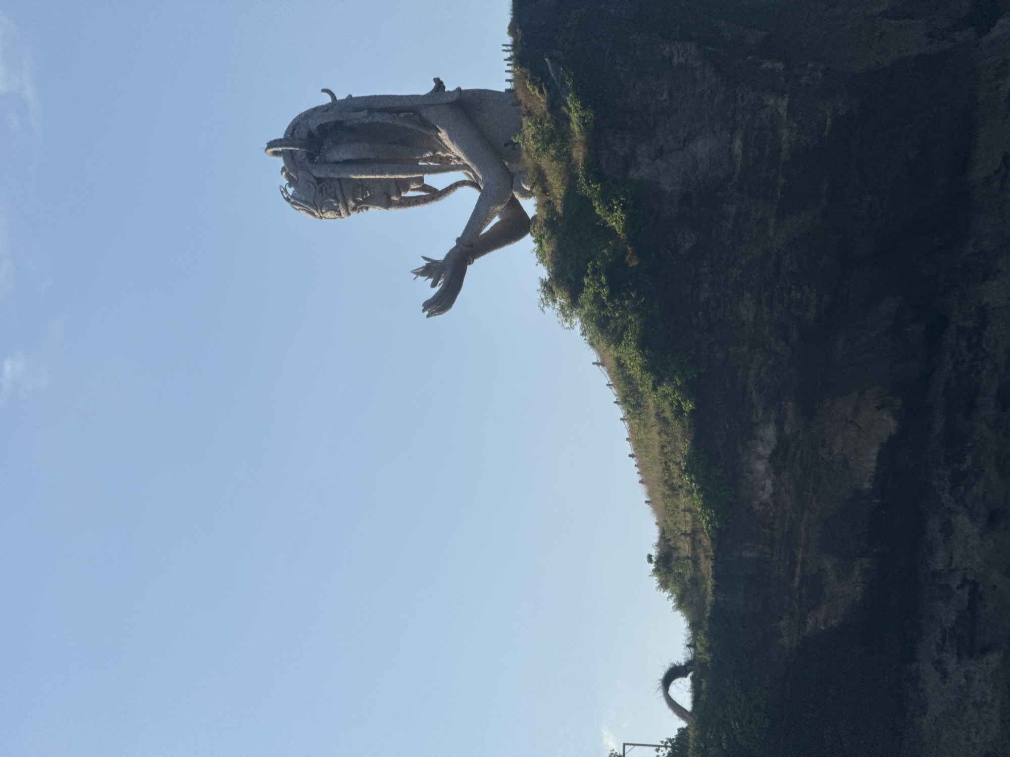 The image shows a large stone statue of an individual in a crouching position, located on a cliffside. The statue is heavily detailed and has what appears to be intricately designed headgear. The figure's arms are crossed, and its hands are visible. The cliff is covered in vegetation, including green foliage. In the background, there is a clear blue sky. The statue is located in Nusa Penida, Indonesia.