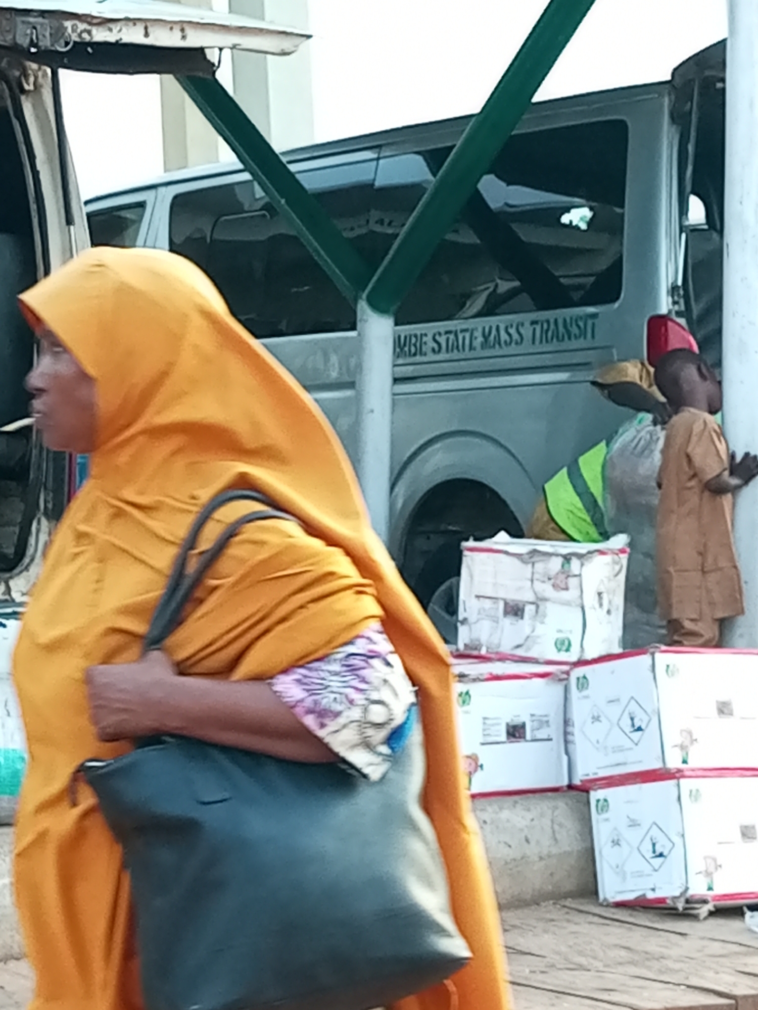 In Gombe, Nigeria, a woman wearing a bright orange hijab and a patterned blouse is seen from the side, carrying a dark-colored tote bag over her left shoulder. She is standing near the open back of a vehicle, possibly a van, which has "GOMBE STATE MASS TRANSIT" printed on its side. Behind her, a young boy in a tan outfit with a red cap is standing next to a pile of white boxes with red trim. Some of these boxes display hazard symbols, including a biohazard warning. Other individuals, including one wearing a high-visibility vest, are partially visible in the background, suggesting activity related to loading or unloading goods. The ground is a textured surface, likely concrete or asphalt, and the overall scene appears to be outdoors, possibly at a transportation hub or market.