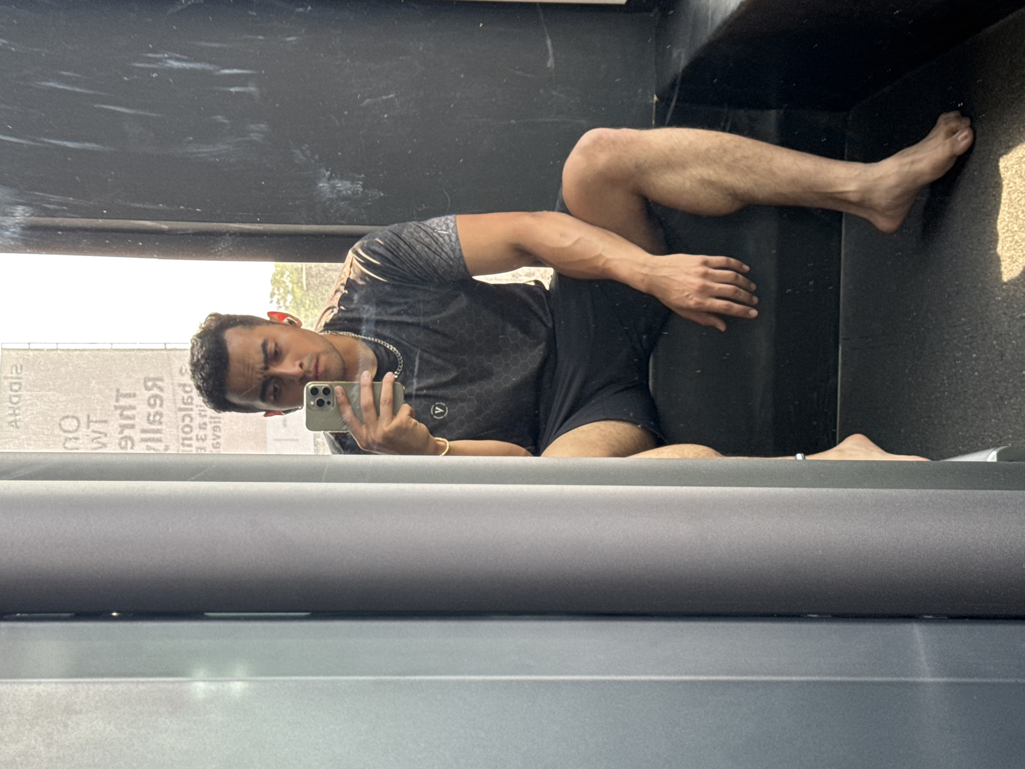 A man is reflected in a mirror, likely in a gym setting. The man, possibly of Indian descent, is wearing a patterned black t-shirt and black shorts. He is holding a smartphone, seemingly taking a photo of himself in the mirror. He has a gold bracelet on his left wrist. The mirror appears to be in a gym setting, with a gray and black backdrop. The scene implies it may be in Kolkata based on the user's instructions, but that can't be certain from the image alone. The light suggests it is daytime. There's signage or writing visible in the mirror's reflection, possibly an advertisement or informational material. The man is partly reclined, with one leg extended along the black surface in the background.