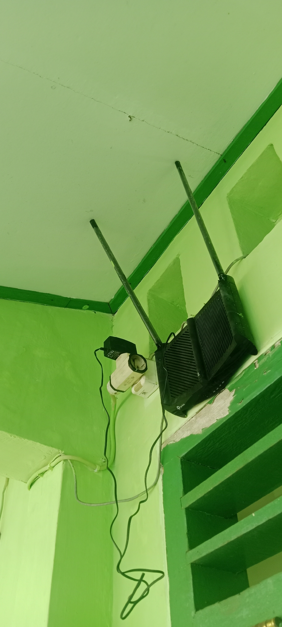 An interior shot shows a black TP-Link Wi-Fi router mounted on a light green wall with two antennas. To the left of the router, a white electrical outlet is visible, with a black power adapter plugged into it, and a white cylindrical object with what appears to be a light sensor attached to the adapter. The wall has some chipped paint. Above and to the left of the router, a crack is visible in the light green ceiling. To the right of the router, part of a green-painted louvered window frame is visible. The image appears to be taken in a residential or small business setting in Tuban, Indonesia.