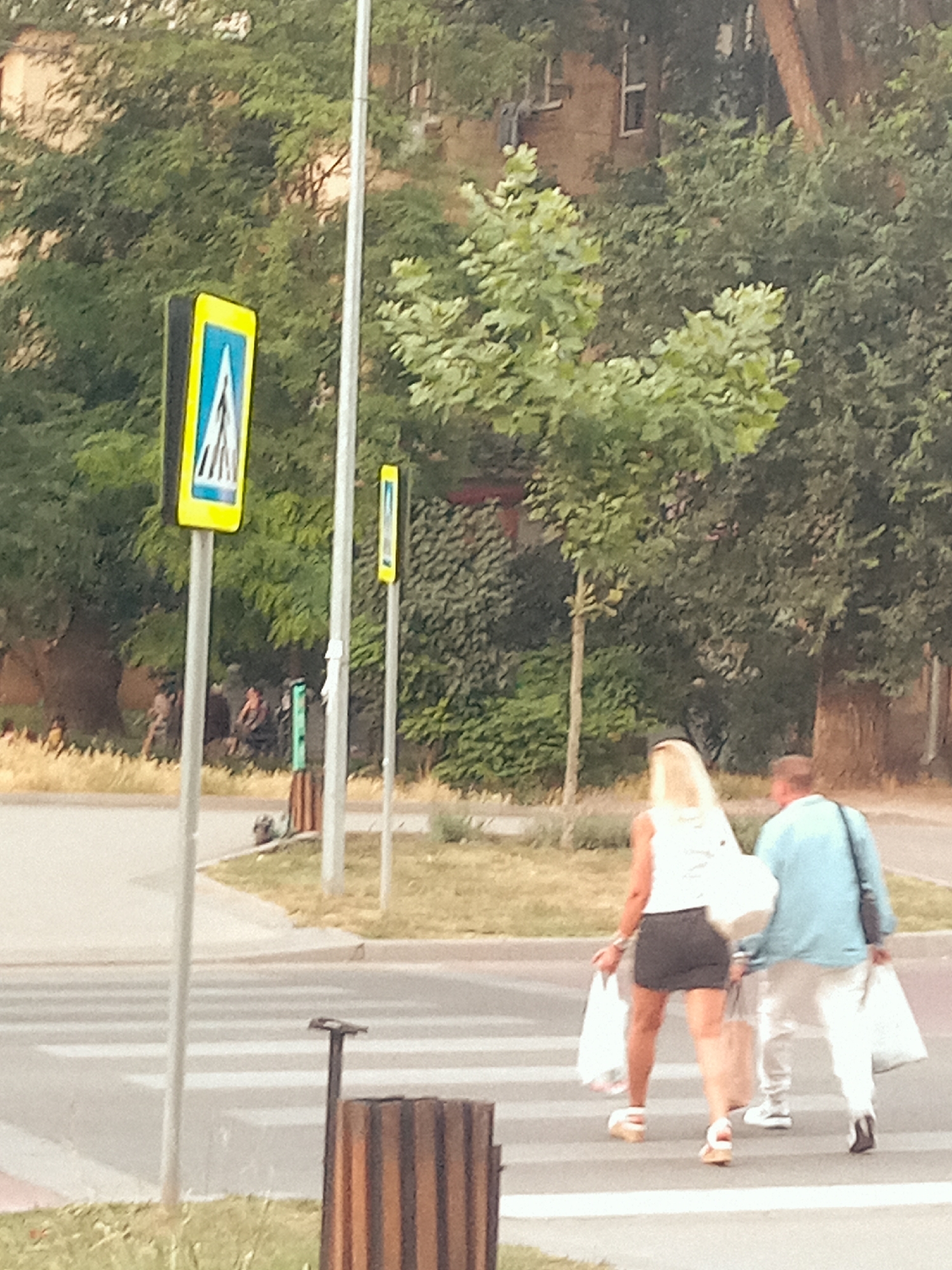 The image captures a street scene with a pedestrian crossing in the foreground. Two individuals, a woman with blonde hair wearing a white top and a black skirt, and a man in a light blue shirt and white pants, are walking across the zebra stripes, each carrying white shopping bags. The woman is slightly ahead of the man. Behind them, a yellow pedestrian crossing sign with a blue pictogram of people walking is visible, mounted on a pole. Another smaller yellow sign is also present further down the road. The background consists of trees and some buildings, suggesting an urban or suburban environment. The lighting indicates it is daytime, possibly late afternoon or early evening. The overall mood appears to be casual, with the individuals likely returning from shopping. The image is slightly blurry, contributing to a sense of candidness. There is no visible text in the image besides the symbols on the traffic signs.