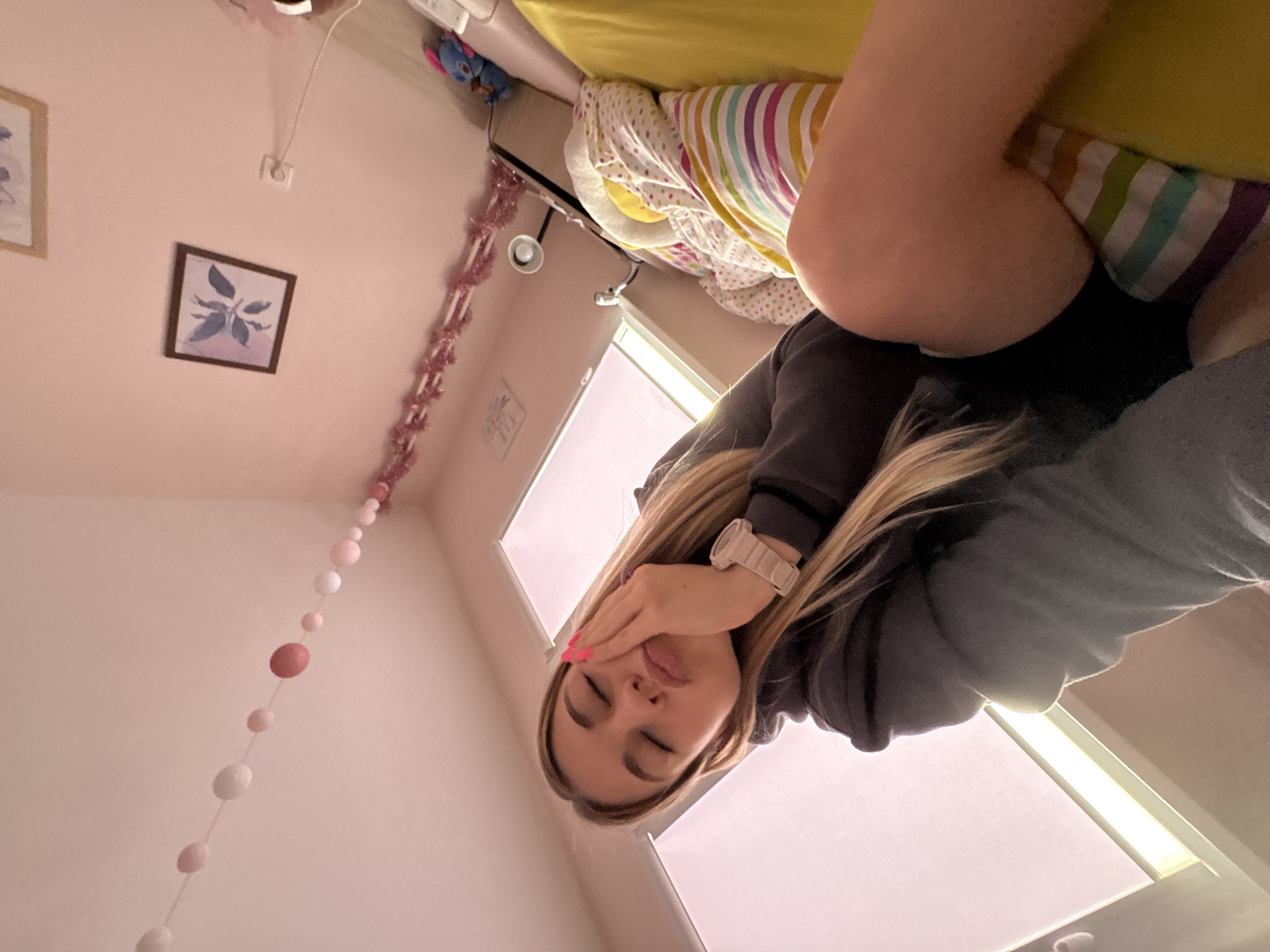 The image captures a close-up, slightly upward-angled view of a young woman resting or relaxing in what appears to be a bedroom. She has long, straight blonde hair and is seen from the chest up, with her eyes closed or partially closed. Her left hand, adorned with bright pink nail polish, rests gently on her cheek and temple. She is wearing a dark grey or black hoodie and a light-colored wristwatch on her left wrist. Her expression conveys a sense of tiredness, sleepiness, or deep contemplation.
The setting is a pastel pink room, likely a bedroom, with various decorative elements. In the background, a bed is visible with a vibrant yellow sheet or blanket and a striped pillow featuring colors like yellow, orange, white, green, blue, and purple. A white comforter with multi-colored dots is also on the bed. A small blue plush toy is positioned near the head of the bed. The wall features two framed pictures; one clearly shows light purple/blue leaves against a white background in a dark frame. Decorative strings, including a garland of pink tinsel and a string of varying shades of pink and white spherical beads, hang vertically along the wall. A white electrical outlet and a small ceiling light fixture with a wire are also visible. Behind the woman, a rectangular window or light panel provides ambient daylight. No visible text is present in the image. The overall atmosphere is personal and quiet, suggesting a moment of rest.