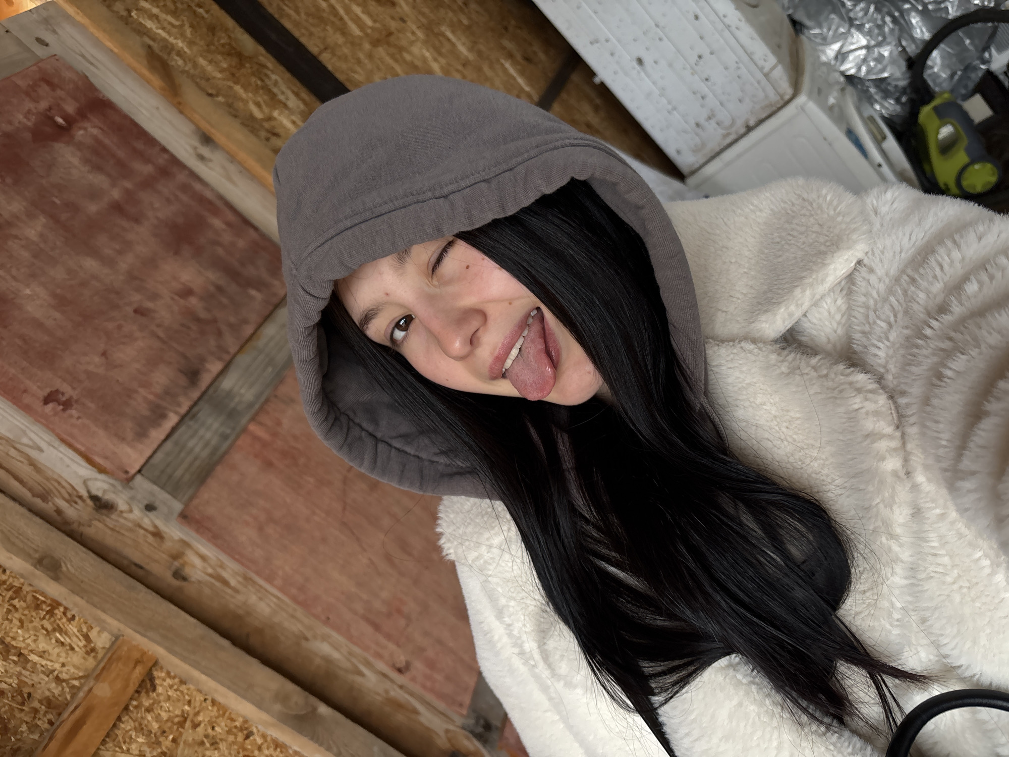 A young woman with long, dark hair and fair skin is wearing a gray hoodie and a white, fleece-like jacket. She is sticking her tongue out and winking. The background consists of wooden planks and a white object, suggesting a garage or shed-like environment. There is no location information available to reference Фоки, Russia.