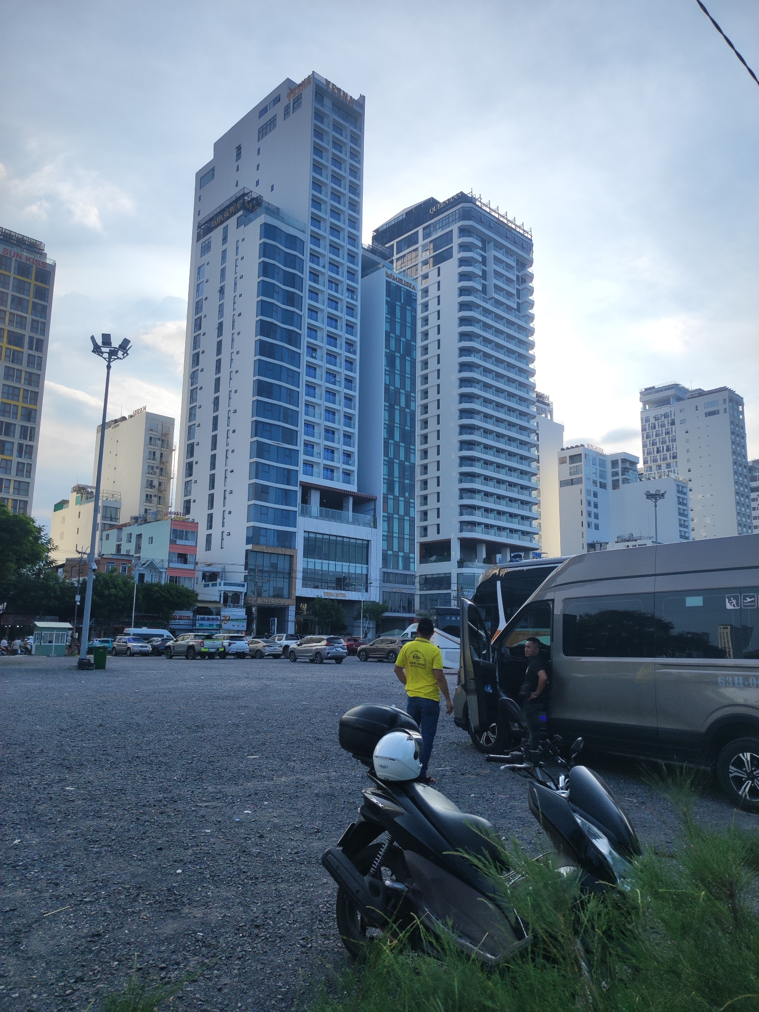 The image depicts an outdoor scene in Nha Trang Ward, Vietnam, with a focus on a parking lot and several modern high-rise buildings. In the foreground, two motorcycles are parked. One is a black scooter with a white helmet resting on its seat, partially obscured by green foliage. The second motorcycle is also black and parked nearby.
In the mid-ground, a tan van is parked with its side door open, and two men are interacting near it. One man, wearing a yellow t-shirt and blue jeans, stands outside the van, facing away from the camera. The other man, wearing a dark t-shirt and shorts, is seated inside the van, his arm resting on the open door. Several other vehicles, including cars and SUVs, are parked in the lot behind the van, with some appearing to be facing a street.
The background is dominated by the imposing architecture of several tall, modern buildings. These structures feature repetitive window patterns and contemporary designs. A tall lamppost stands to the left of the frame. The sky above is partly cloudy, suggesting either early morning or late afternoon light. The ground of the parking lot is covered with gravel. The overall impression is of a busy urban environment with commercial and residential structures.