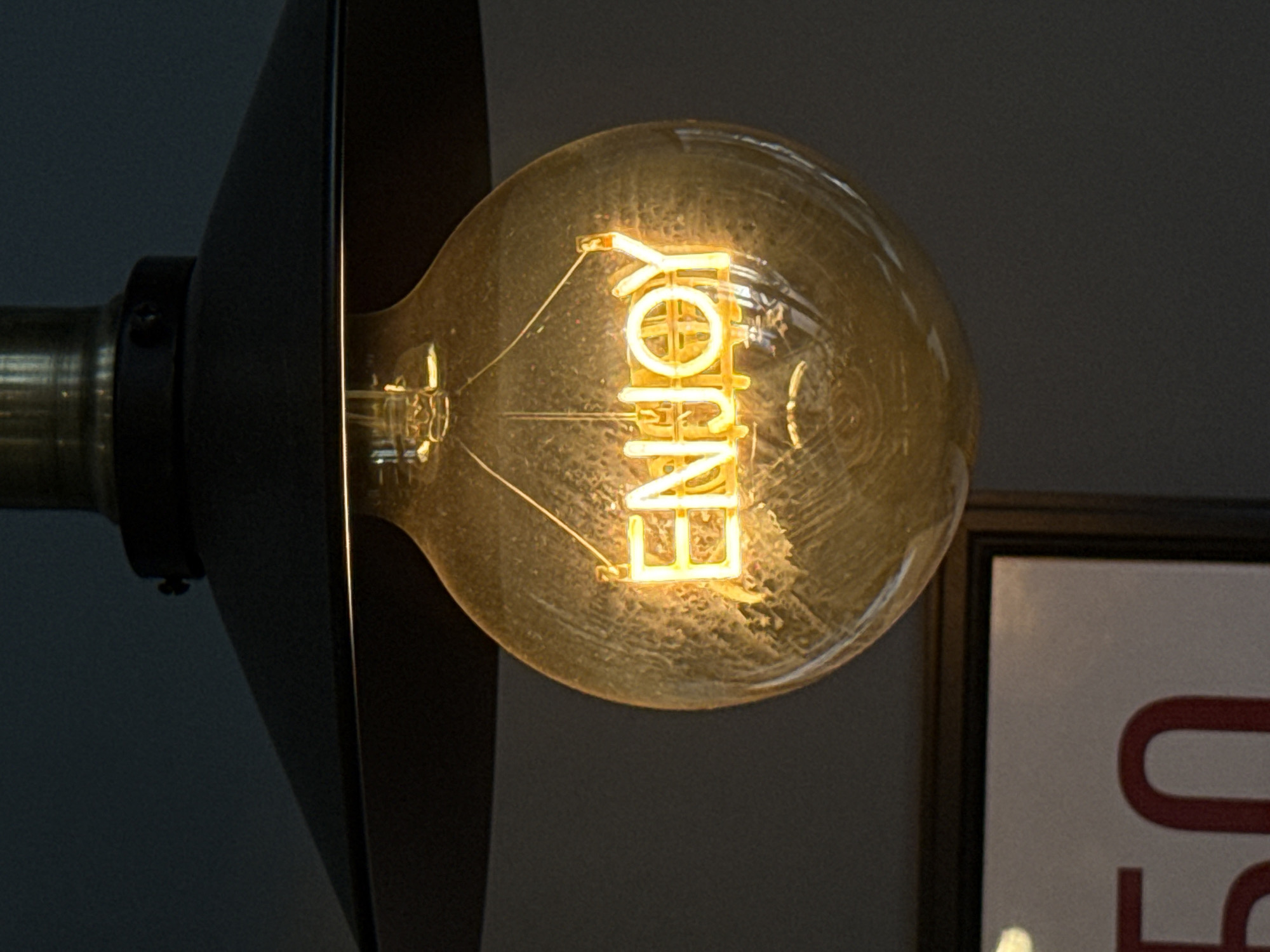 A close-up, vertically oriented photograph captures a decorative light bulb, illuminated and casting a warm, golden glow. The main subject is a large, clear glass bulb, inside which the word "ENJOY" is formed by intricate, glowing LED filaments. The lettering is bright yellow-orange against the darker interior of the bulb.
The bulb is attached to a light fixture, with a dark, possibly brass-colored, cylindrical base visible on the far left. A portion of a black, cone-shaped light shade extends from the base, partially obscuring the left side of the bulb and the background. The background throughout the image is a dark, plain surface, likely a wall, which creates a strong contrast with the bright light source.
To the right of the light bulb, a framed object is partially visible. It shows a segment of a white background with a large, bold red character, which appears to be part of a letter like "U" or "O".
The image depicts an indoor scene, likely in a commercial establishment such as a cafe, restaurant, or shop, given the decorative nature of the bulb and the visible sign fragment. The illumination of the bulb suggests the surrounding environment may be dimly lit, indicating either evening/night or a deliberately atmospheric interior. The scene is set in Krasnoyarsk, Russia.