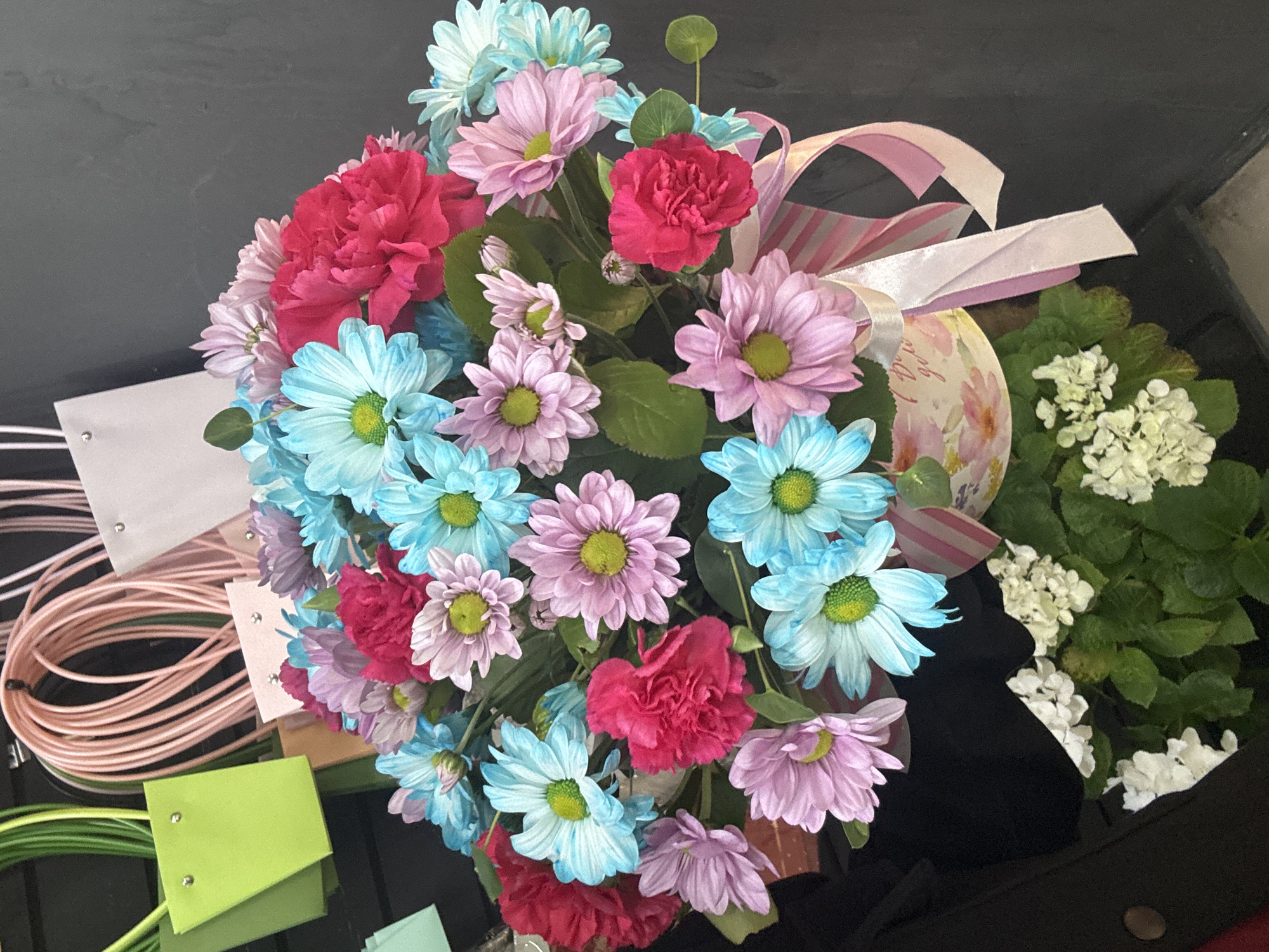 A colorful bouquet of chrysanthemums and carnations is presented in a decorative pot adorned with a pink and white striped ribbon. The flowers are arranged in a dense display, featuring vibrant shades of pink, blue, and fuchsia. Some of the chrysanthemums have pale green centers, and their petals are tinged with white. The carnations, with their ruffled petals, add texture and depth to the arrangement. The pot itself has a floral pattern and appears to be made of paper or cardstock. In the background, there are several gift bags in pastel colors, including lime green, pale pink, and white, some with rope handles. The scene is set against a dark, textured wall. Below the bouquet, a cluster of white hydrangeas and green foliage is visible. The overall impression is that of a festive floral display, likely intended as a gift.