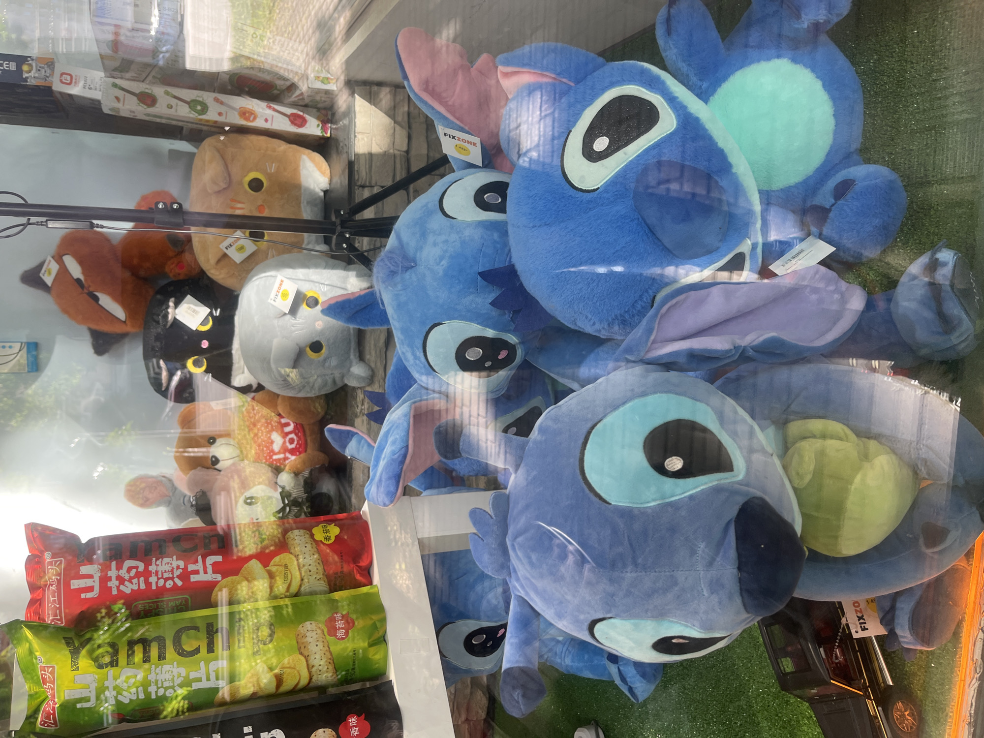 The image is a close-up, eye-level shot of a claw machine filled with plush toys and snacks. The primary subjects are several large, blue Stitch plush toys from Disney's Lilo & Stitch, piled on top of each other. To the left of the Stitch plushies are other plush toys, including a brown cat with large yellow eyes, a black cat with yellow eyes, and a grey cat with yellow eyes. There are also some snacks visible at the bottom of the machine. These snacks are packaged in red and green bags, with Chinese text and images of potato chips or yam slices on them. One bag clearly reads "Yamchip" and "YAM SLICES". The claw mechanism of the machine is partially visible, suspended above the toys. The background appears to be a brick wall. Reflections are visible on the glass surface of the machine, obscuring some details. The overall scene suggests a retail or entertainment setting, likely an arcade or shopping mall. Visible text includes "汇江码头", "山药薄片", "Yamchip", "YAM SLICES", "海苔味", "番茄味", and "FIXZONE" which appears on tags attached to some of the plush toys. The lighting suggests it is daytime, possibly indoors under artificial lights. No people are visible in the frame.