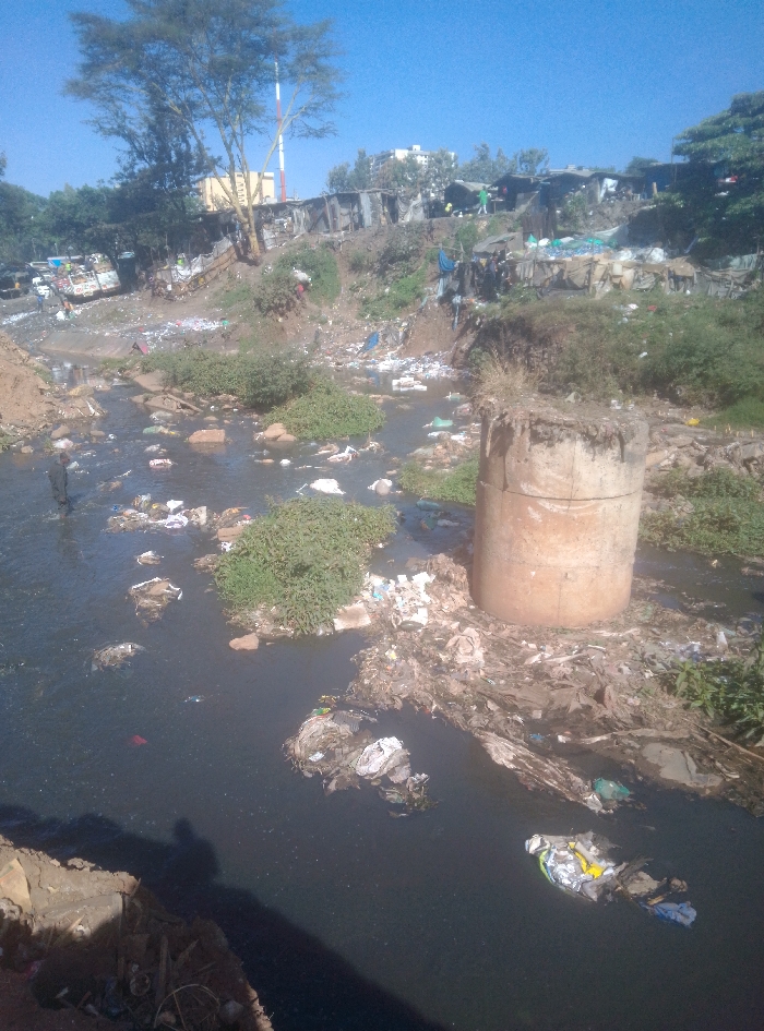 The image depicts a severely polluted river flowing through an informal settlement in Nairobi, Kenya. The river's water is dark and murky, extensively covered with floating refuse including plastic bottles, bags, fabric, and various discarded materials, forming dense accumulations throughout the visible expanse. A prominent, approximately 2-meter-tall cylindrical concrete pillar stands in the mid-stream on the right side, serving as a collection point for a large mound of debris.
The riverbanks are steep, eroded, and covered with dirt and scattered trash. Numerous informal dwellings, constructed primarily from corrugated metal sheets, wood, and other salvaged materials, are densely packed along the upper embankments, indicative of a slum environment. Several individuals are visible on the embankments and among the structures, engaged in unidentifiable activities.
Sparse green vegetation, including bushes and small plants, grows along the banks and in isolated patches within the river. A large, deciduous tree with light-colored bark stands on the left bank against a clear blue sky. In the upper background, a red and white striped tower or mast is visible, with additional informal structures and some taller buildings in the distant background. The scene is illuminated by bright sunlight, casting shadows.