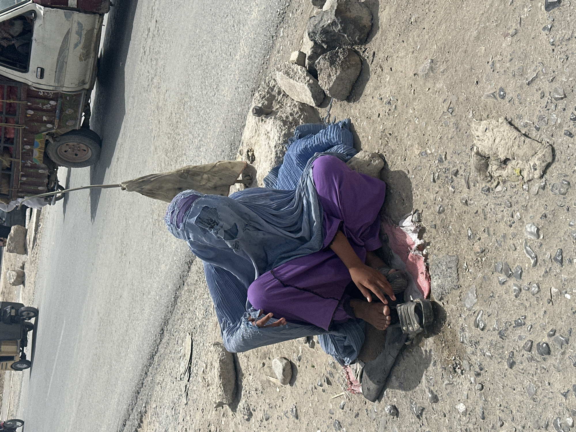 A person is seated on the ground near a road in Quetta City Tehsil, Pakistan. They are wearing a light blue burqa and purple clothing. The person's bare feet are visible. A gray shoe sits nearby. The road has vehicles on it. The ground is dirt and gravel with some rocks.