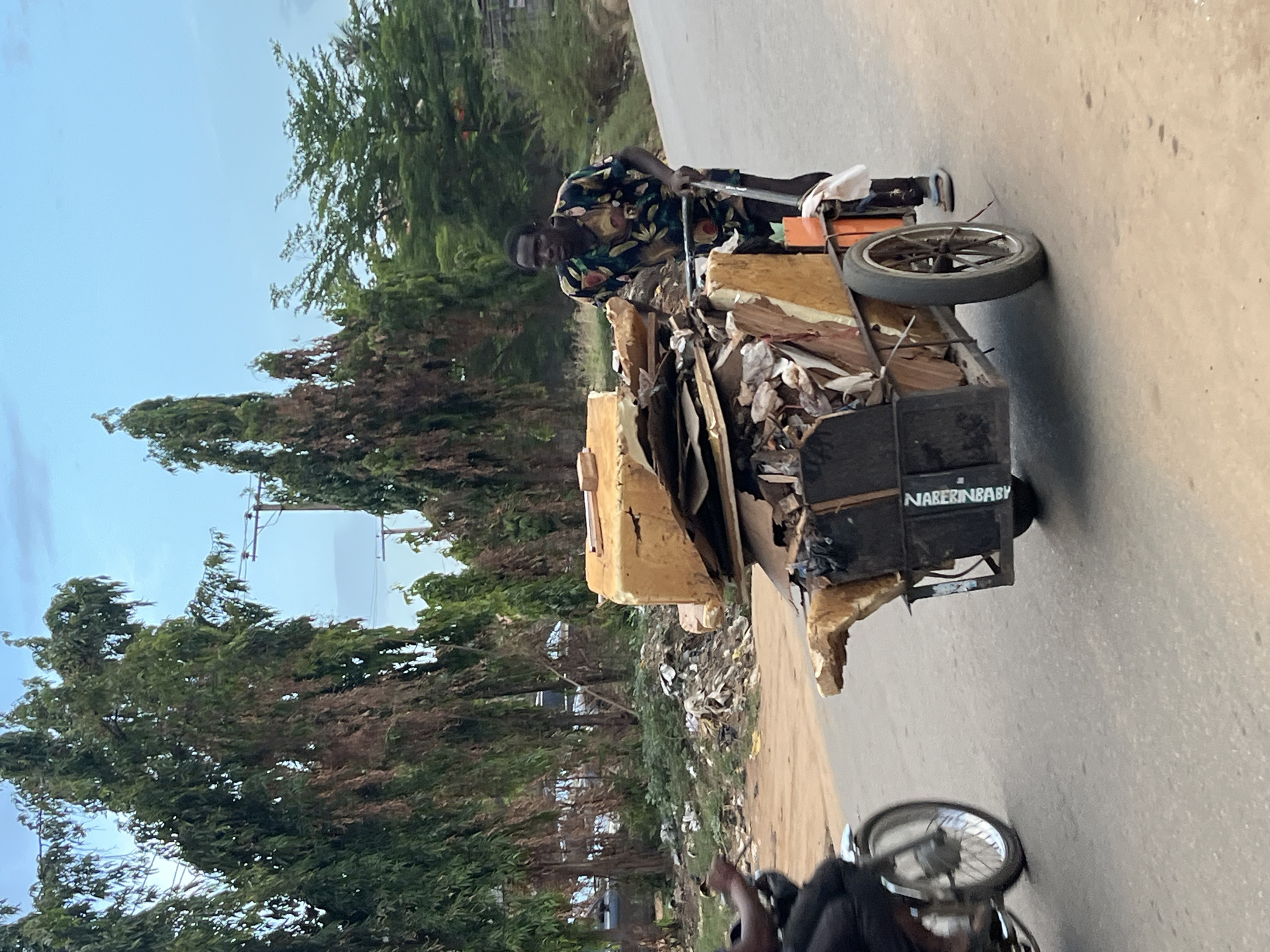 The image, taken in Abuja, Nigeria, depicts an outdoor scene under bright daylight. A man is the central subject, standing next to a large, two-wheeled cart filled with various discarded materials such as cardboard, paper, plastic, and wood scraps. He is dark-skinned, wearing a vibrant multi-colored (green, yellow, and black) patterned shirt and dark shorts, and is looking directly towards the camera with a neutral expression. He appears to be pushing or managing the cart.
The cart is robustly constructed, made of wood and metal, with the text "NABIRINBABY" visible in white letters on its black metal side panel. The scene is set on a paved road, with a blurred wheel of another vehicle partially visible in the immediate foreground, suggesting movement or another observer. Lush green trees line both sides of the road under a light blue sky with minimal cloud cover. The ground adjacent to the road shows evidence of litter and debris, consistent with a roadside environment where waste might accumulate. The overall activity suggests waste collection or scavenging.