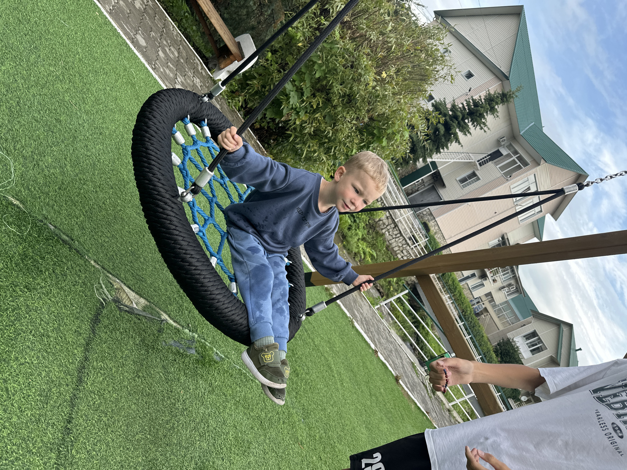 The image depicts a young Caucasian boy with blonde hair, wearing a blue sweatshirt and blue pants, sitting on a black and blue rope swing. He is looking to his right with his mouth slightly open. His hands are gripping the ropes of the swing. In the background, a modern building with a green roof and light-colored siding is visible. There is also a lush green tree next to the building. The foreground shows a green artificial turf surface, with a section appearing to be damaged or torn. To the right of the frame, a person's arm and torso are visible, wearing a white t-shirt and black shorts with the number "29" on them. This person is holding a beaded bracelet. The overall scene suggests an outdoor recreational area, possibly a backyard or playground, in a suburban or semi-rural setting. The location is specified as Danilovka (Altai Krai), Russia.