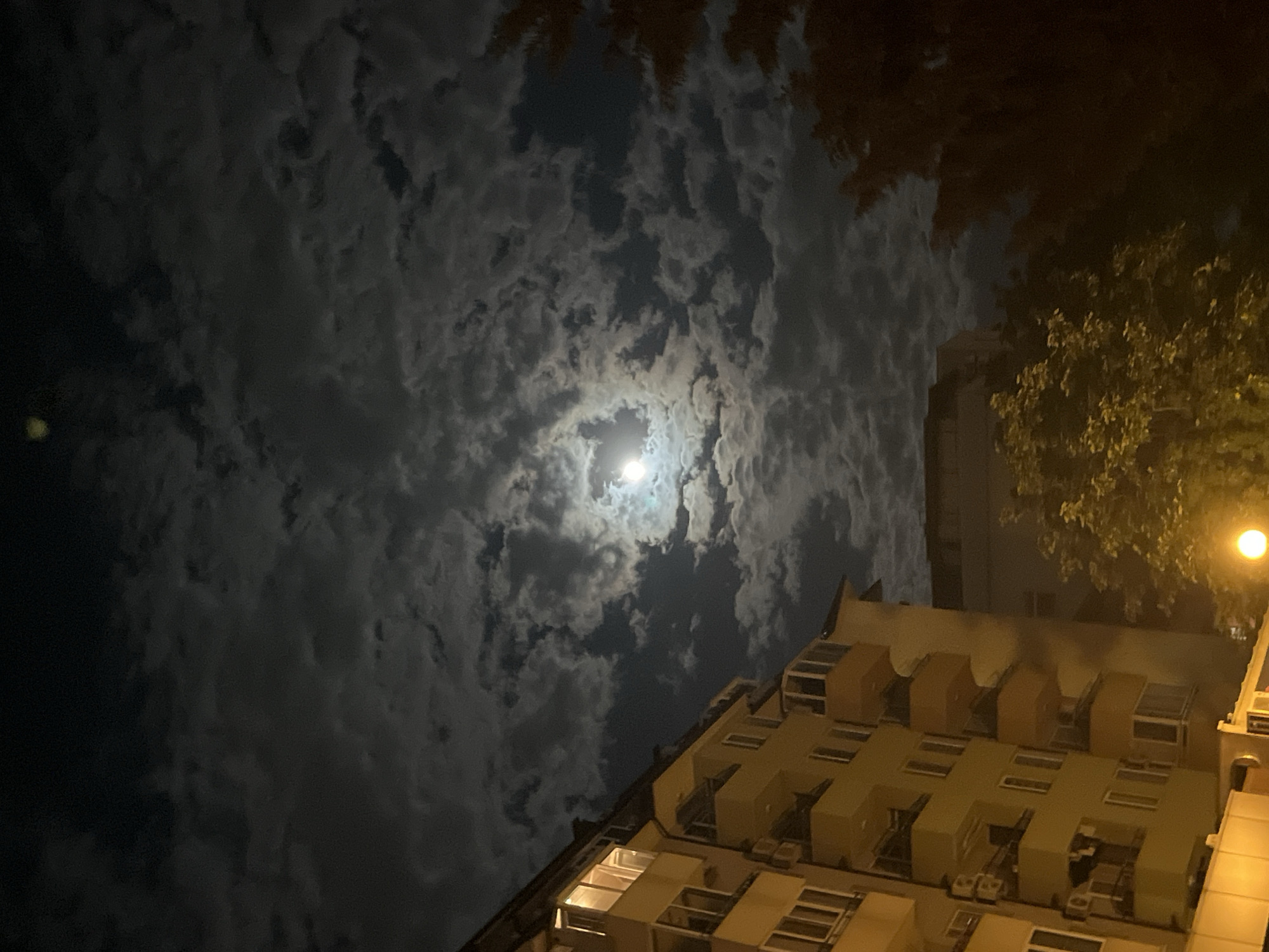 The image depicts a night scene. In the center of the sky, there is a bright moon surrounded by clouds. At the bottom right of the frame is a light-colored building with multiple windows and protrusions. A street light is shining near the top right, with tree branches near it. The location is Unknown, Unknown.