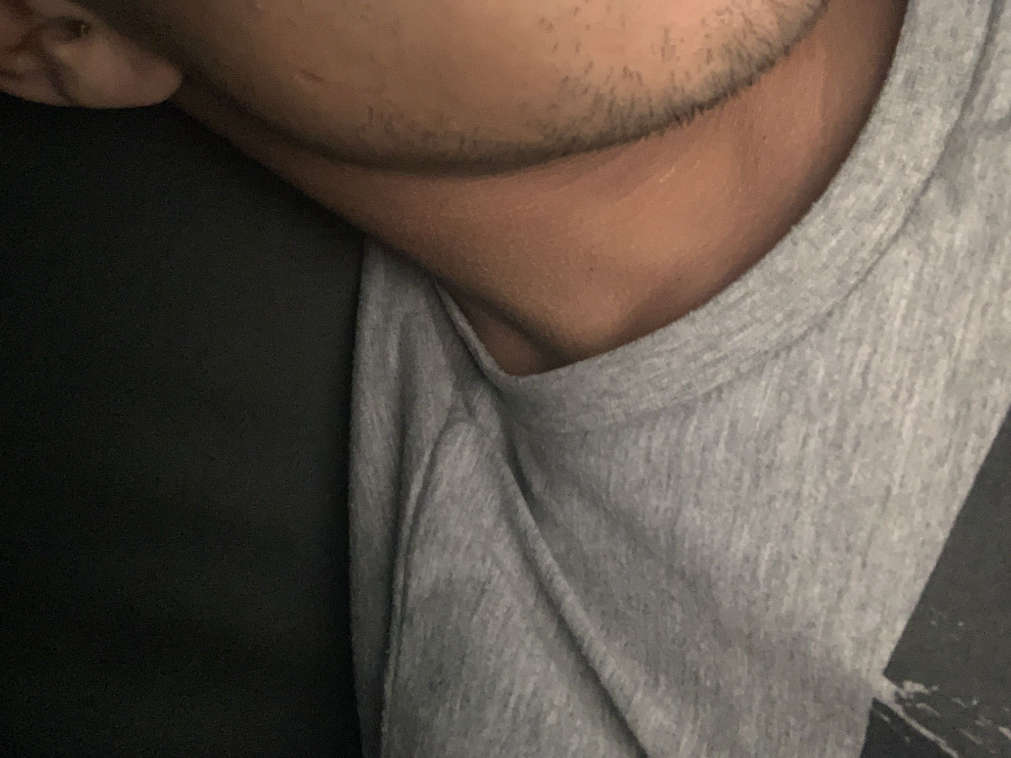 The image shows a close-up view of a person's neck, lower face, and shoulder, wearing a heather gray t-shirt. Part of the left side of the image is black, possibly clothing or a dark background. The person has short facial hair. No information is provided in the image or crops suggesting it was taken in Kalitinovo, Bulgaria.