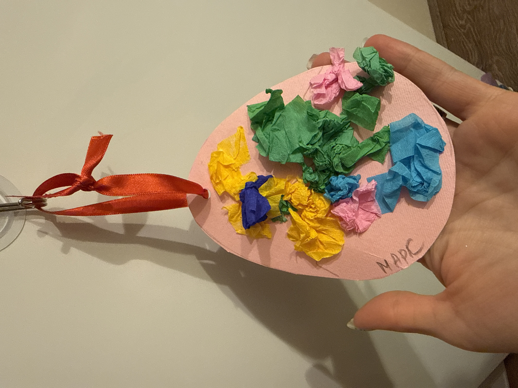 A hand holds a pink egg-shaped ornament decorated with colorful tissue paper. The tissue paper is crinkled and arranged in clusters of yellow, green, blue, and pink. A red ribbon is attached to the egg with a metal clip. The name "MARC" is written on the egg. The background shows a light-colored surface and a partial view of a wall, indicating the setting is indoors. The location of the city of Zelenika, Montenegro, is not discernible.
