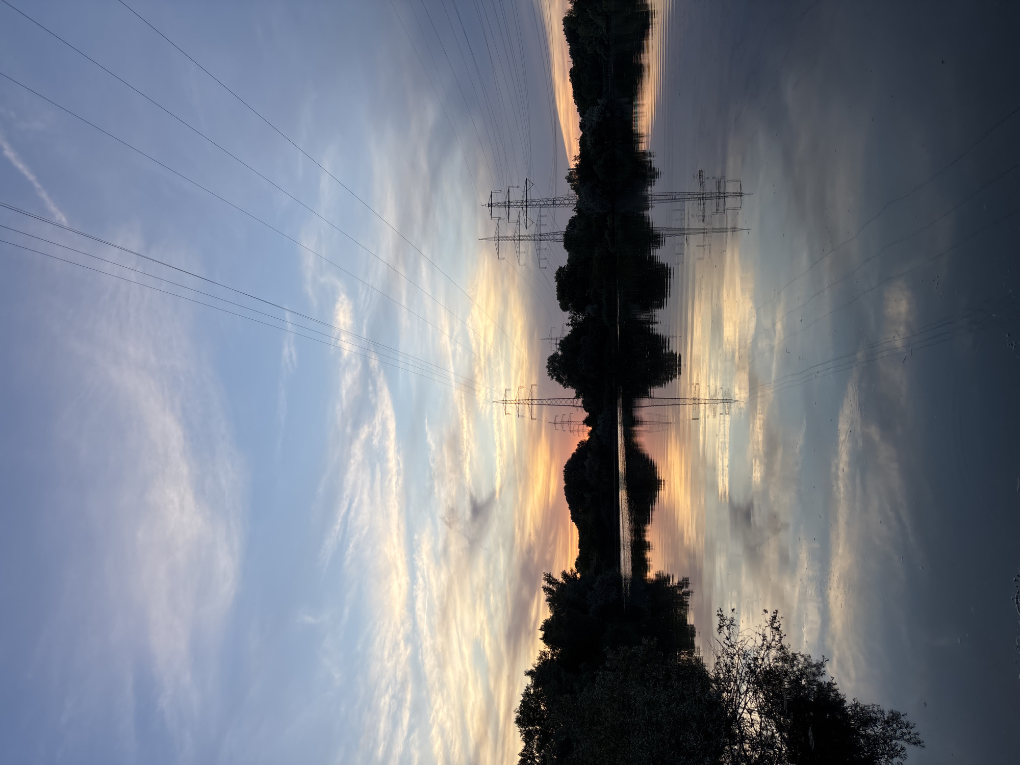 The image depicts a tranquil natural scene at dusk, likely captured in Ludwigshafen am Rhein, Germany, given the presence of pylons and the landscape composition. A body of water occupies the foreground, reflecting the sky and the surrounding environment. The sky is a canvas of soft hues, transitioning from a pale blue at the top to warmer oranges and yellows near the horizon, indicating the setting sun. Wispy clouds are scattered across the sky, adding texture and depth.
Dominating the middle ground are silhouetted trees lining the opposite bank of the water. Emerging from behind the trees are several large electricity pylons, typical of German infrastructure. These structures are starkly outlined against the colorful sky and also mirrored in the calm water. Overhead, power lines stretch across the frame, further emphasizing the man-made elements within the natural setting. The overall atmosphere is one of peaceful twilight, with the natural beauty of the sunset complemented by the industrial presence of the pylons. No people are visible in the image.