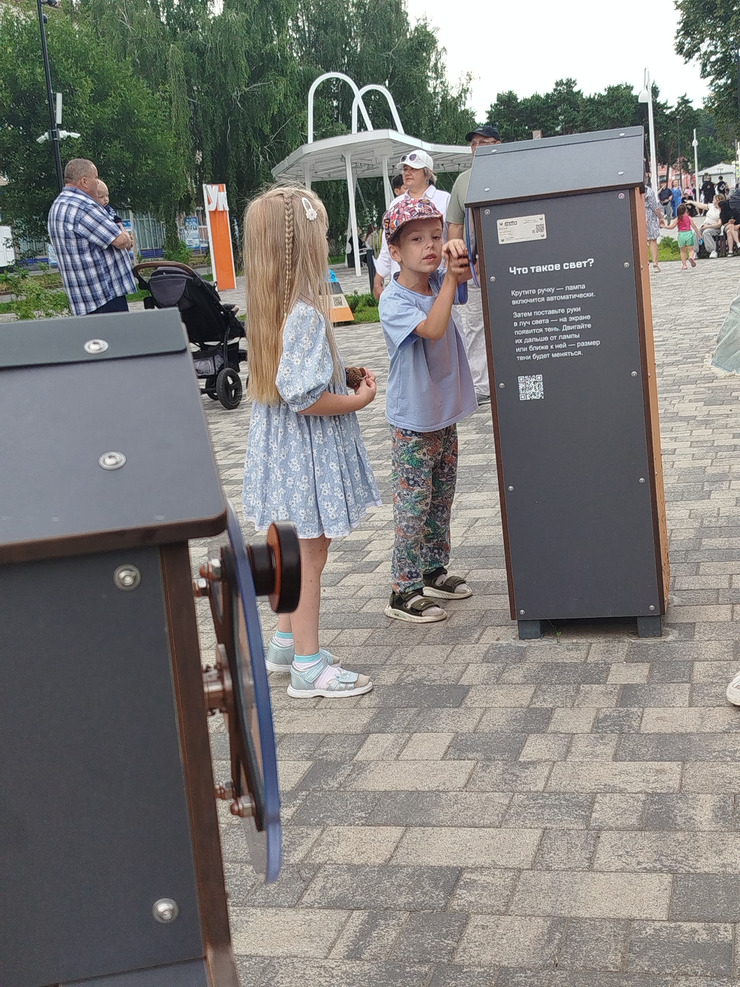 In Zavodoukovsk, Russia, on a cloudy day, two children interact with an educational display in an outdoor public space. A young boy, wearing a patterned cap and a light blue t-shirt with colorful patterned pants, is actively engaged with the display, his hands reaching towards it. Beside him, a young girl with long blonde hair, styled in braids, stands in a blue floral dress. She holds a small object in her hand.
The display itself is a dark-colored, vertical structure with text and a QR code. The text, in Russian, asks "What is light?" and provides instructions for an experiment: "Turn the handle - the lamp will turn on automatically. Then place your hands in the beam of light - a shadow will appear on the screen. Move them further from the lamp or closer to it - the size of the shadow will change."
In the background, other people are visible, including a man holding a baby and a stroller, suggesting a family-friendly environment. The setting appears to be a park or a public square with paved walkways made of interlocking grey and beige bricks. Green trees and foliage form a backdrop, with some modern white structures suggesting a contemporary park design. The overall atmosphere seems relaxed and educational, with children exploring a hands-on science exhibit.