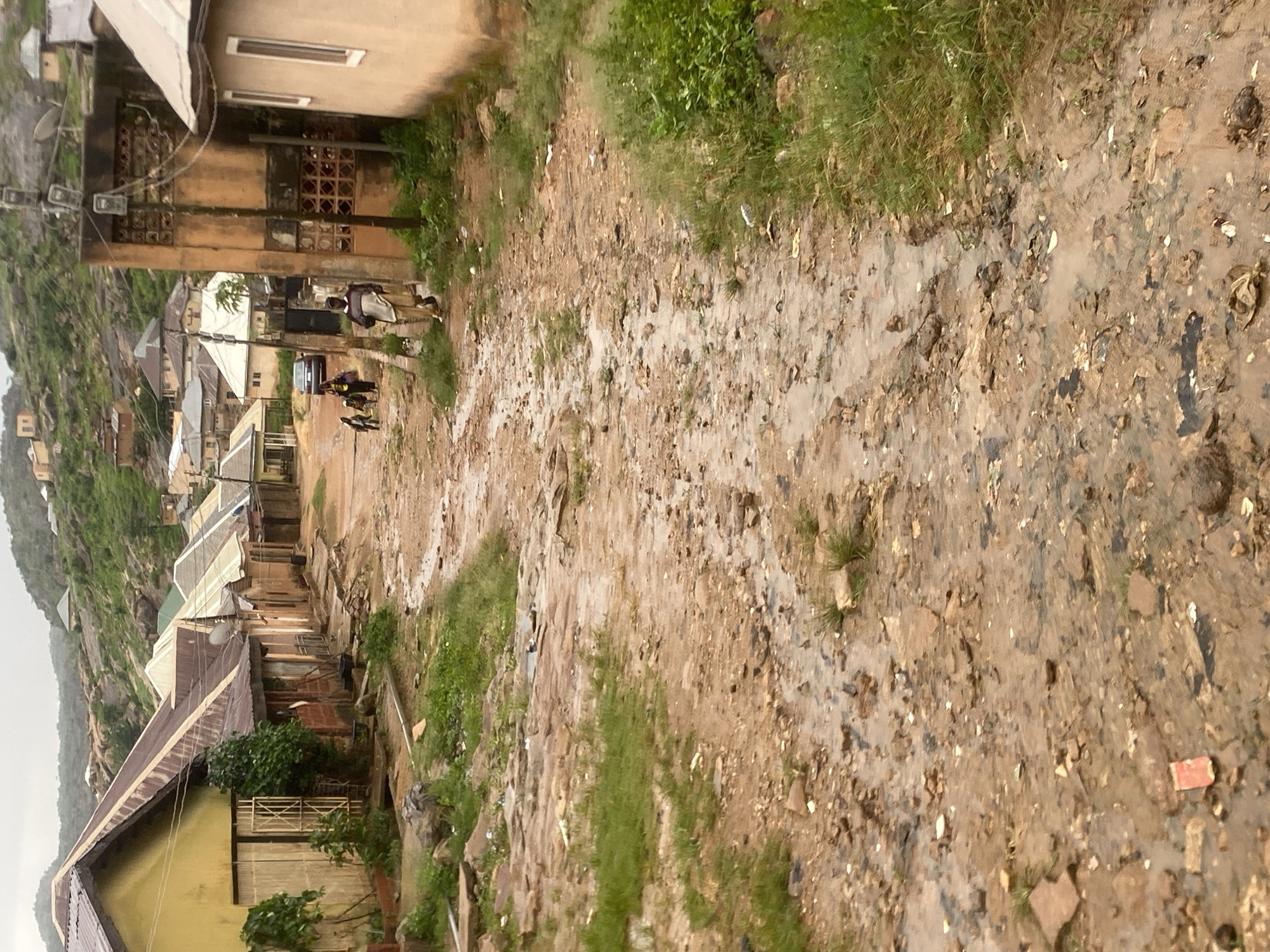 The image depicts a muddy, unpaved street in a residential area of Jos, Nigeria, likely after rainfall. The street is uneven, with puddles and rivulets of water flowing through it, mixed with dirt and debris. On either side of the street, houses are visible, varying in architectural style and condition. Some houses have corrugated metal roofs, while others have lighter-colored roofing materials. Vegetation, including grass and some trees, grows alongside the street and on the slopes of the hills in the background.
In the mid-ground, several people are walking along the street. A man wearing a light-colored tunic and trousers is walking away from the camera. Further down the street, a group of people, including two children, are walking towards the camera. A car is parked on the side of the street in the distance. Utility poles with electrical wires are present, indicating the presence of electricity infrastructure. The overall impression is one of a community in a hilly, somewhat undeveloped urban or peri-urban setting. The scene is characteristic of many areas in Jos, known for its hilly terrain and mixed urban development.