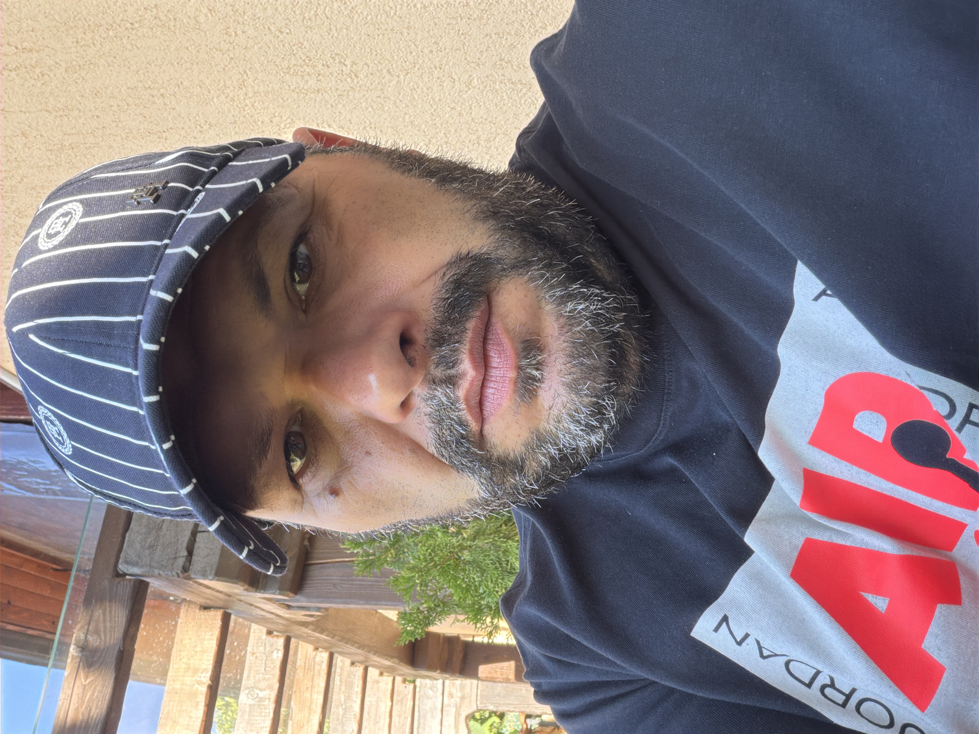 A man with a beard and a striped baseball cap is taking a selfie outdoors. He is wearing a black t-shirt with a graphic design featuring the words "JORDAN AIR". The background shows wooden structures and a beige wall. The image does not contain any information about the location in the city of Smolyan, Bulgaria.