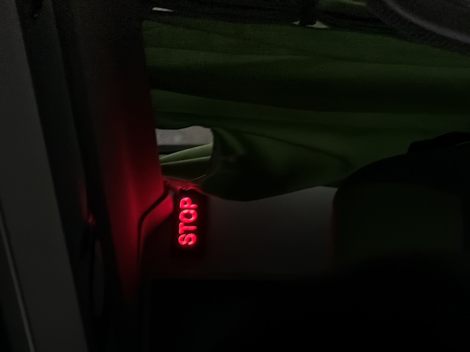 An illuminated red "STOP" sign is visible within the interior of a vehicle located in Moineville, France. The sign, rendered in capital letters, glows brightly against a predominantly dark environment. It is mounted on an overhead panel or compartment. Thin black wiring extends from the top of the sign into the overhead structure. To the right of the sign, a section of a green fabric curtain hangs vertically, partially obscuring a lighter area or window beyond. The foreground and bottom-left portion of the image are obscured by dark, indistinct shapes, consistent with vehicle seating or interior components. No individuals or specific actions are directly observed. The scene is characterized by low ambient light, emphasizing the illuminated "STOP" indicator.