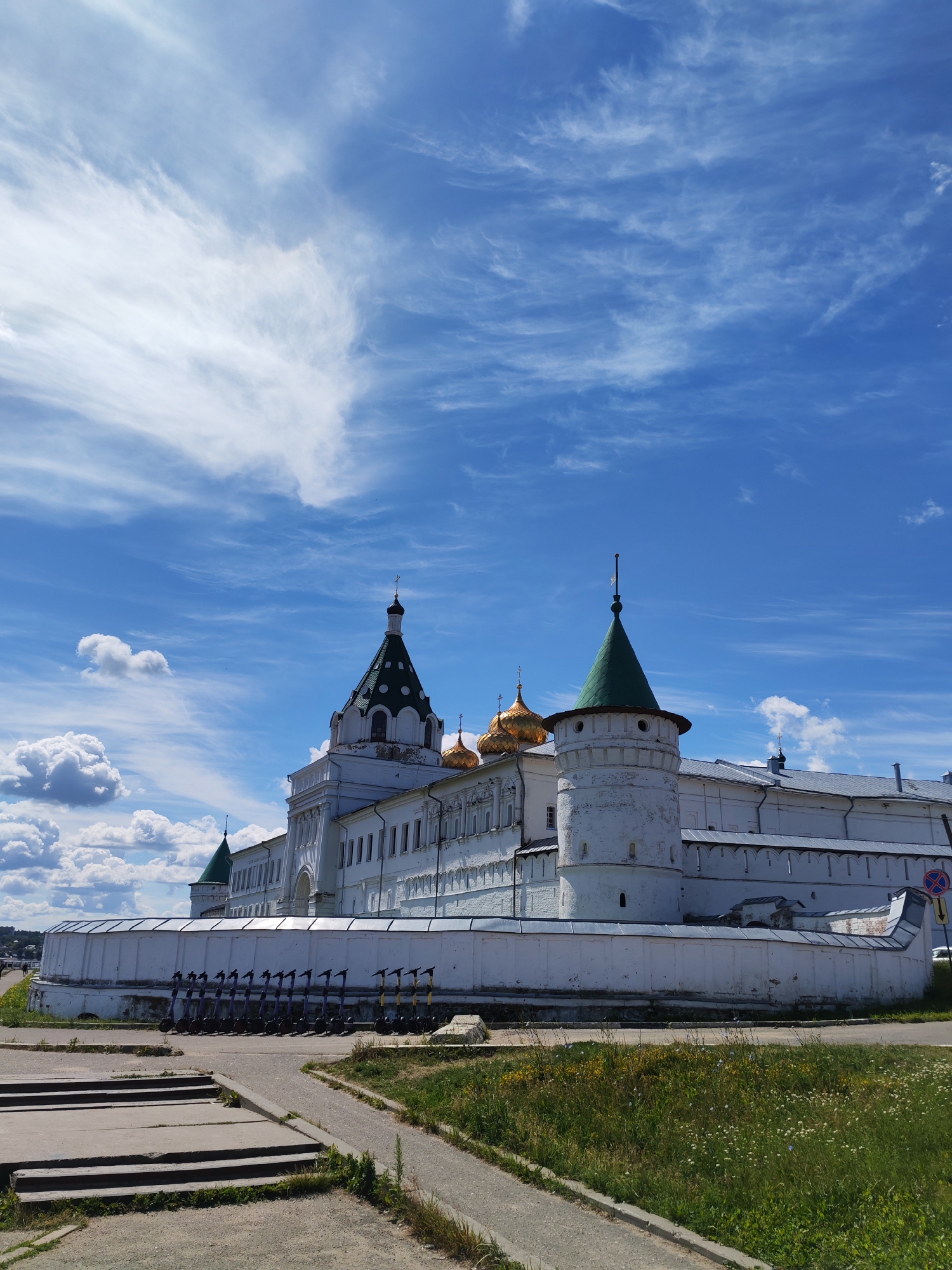 The image depicts the Ipatiev Monastery, a historic Russian Orthodox monastery located on the bank of the Volga River in the city of Kostroma. The monastery features a complex of white buildings with traditional Russian architectural elements, including golden onion domes and green-tiled roofs. A tall, white defensive wall with crenellations surrounds the monastery grounds. In the foreground, a paved walkway leads towards the monastery, with a set of concrete steps to the left and a grassy area to the right dotted with wildflowers. A row of parked electric scooters is visible along the inner side of the wall. The sky above is a vibrant blue with scattered white clouds and wispy cirrus formations, suggesting a clear, sunny day. The scene is devoid of any visible people or animals.