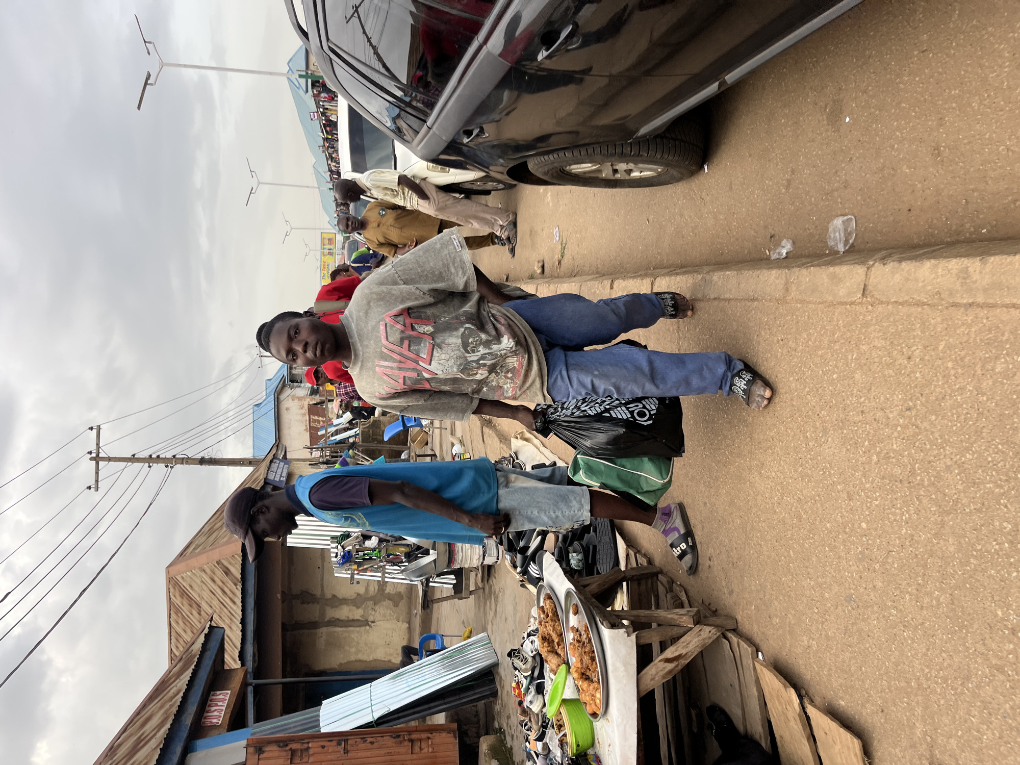 In Bukuru, Nigeria, a street market scene unfolds under an overcast sky. In the foreground, a young man wearing a grey t-shirt with a graphic print and blue jeans walks towards the viewer, carrying two plastic bags, one black and one green. To his left, another man in a blue vest and denim shorts stands near a table displaying platters of fried chicken and various shoes. Behind them, a row of market stalls with corrugated metal roofs lines the dusty street. Several vehicles, including a black SUV, are parked along the roadside. Other individuals are visible in the background, suggesting a busy marketplace. Electrical poles with overhead wires are present, and streetlights are mounted on tall poles. The ground is a mix of dirt and pavement, with some scattered debris.