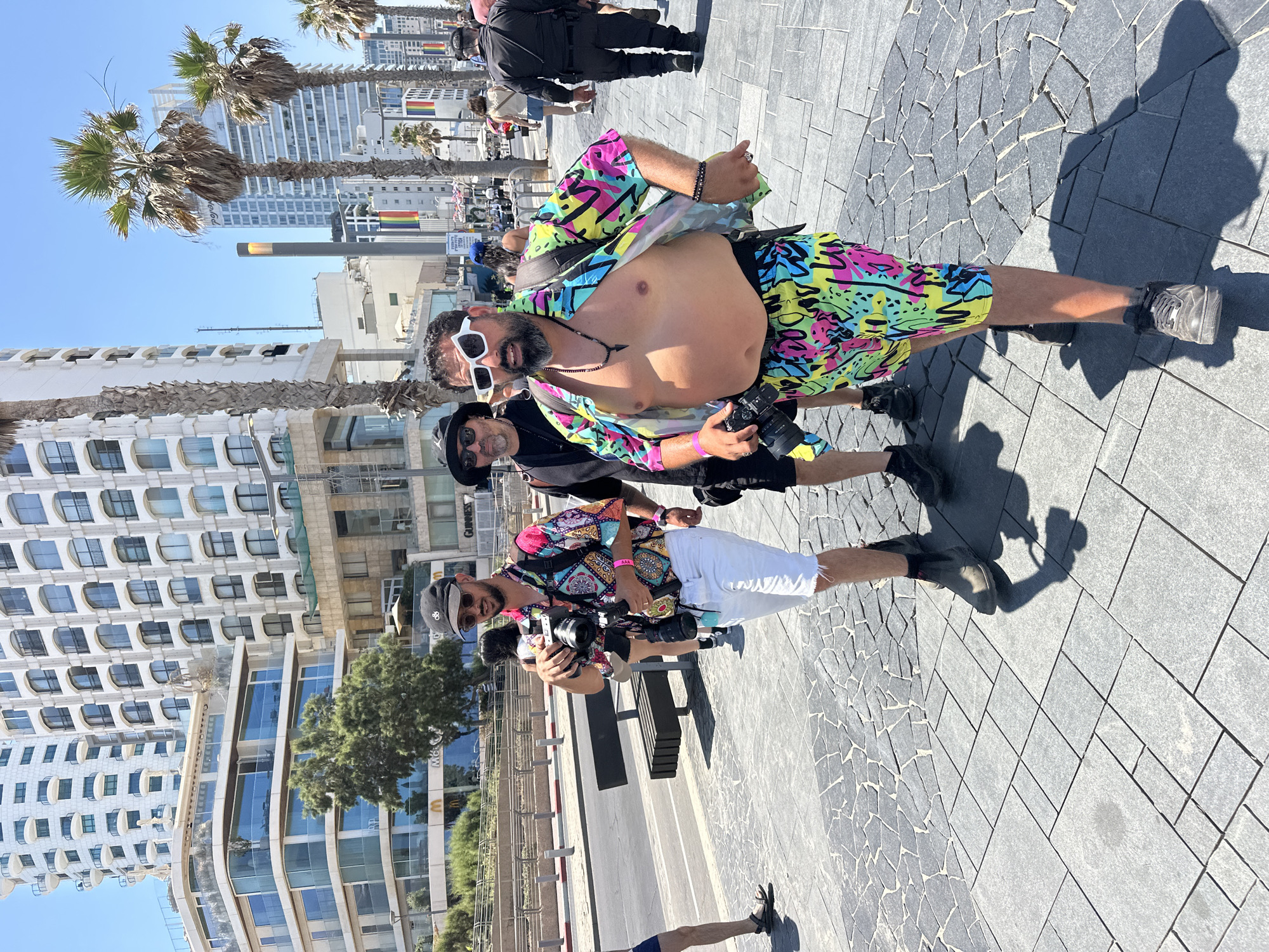 Three men are walking on a tiled sidewalk in Tel-Aviv, Israel, on a sunny day. The man in the foreground, shirtless and wearing brightly colored swim trunks and a matching open shirt with a psychedelic pattern, is holding a camera and looking towards the viewer. He has a beard and is wearing white cat-eye sunglasses and several necklaces. To his left, a man in a black hat, sunglasses, and a black shirt is also holding a camera and walking. Behind him, a third man in a patterned shirt, white shorts, and dark boots is holding a camera. In the background, tall buildings, palm trees, and a busy street with traffic and pedestrians are visible. The presence of rainbow flags on buildings suggests this may be during an LGBTQ+ pride event.