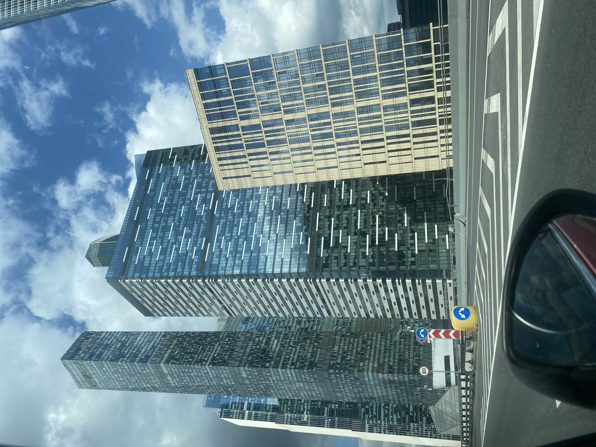 The image depicts a modern cityscape in Moscow, Russia, likely from the perspective of a vehicle on a highway or elevated road. Dominating the scene are several contemporary skyscrapers with glass facades, reflecting the cloudy sky. The buildings vary in height and architectural style, with some featuring intricate grid patterns and others having a more monolithic appearance. In the foreground, the edge of a roadway is visible, including lane markings and a portion of a car's side mirror. Traffic signs, including a speed limit sign of "80" and directional signs, are present along the roadside. A yellow traffic barrel with blue directional arrows is also visible. The sky is partly cloudy with a mix of blue sky and white cumulus clouds.