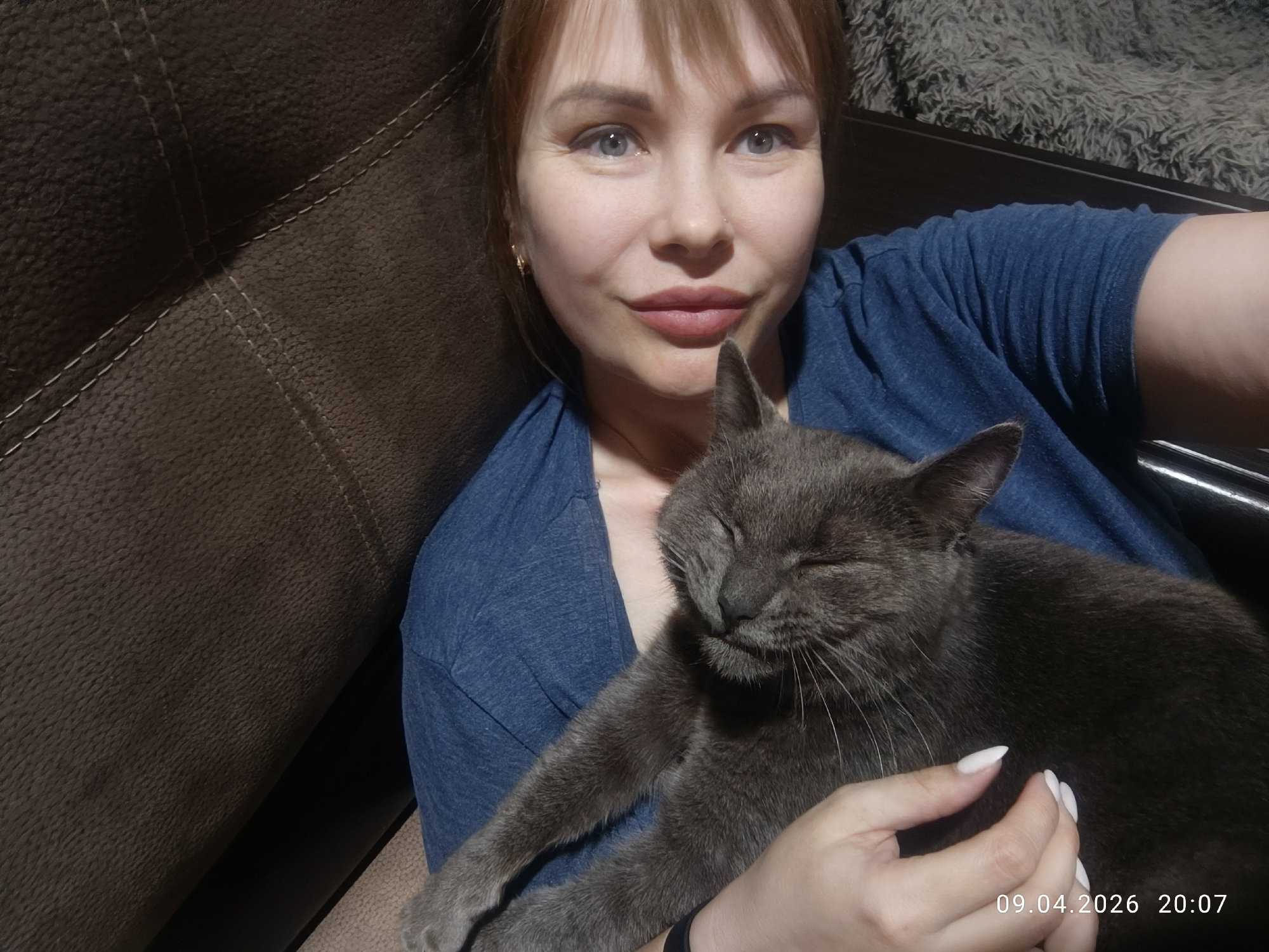 A woman with fair skin, blue eyes, and reddish-blonde hair is holding a gray cat in her arms. She is wearing a blue V-neck shirt. The cat has its eyes closed and appears to be resting. The background includes a brown textured couch and a gray fur-like throw. The date "09.04.2026 20:07" is timestamped in the bottom right corner. The location is Chaikovsky, Russia, based on the timestamp.
