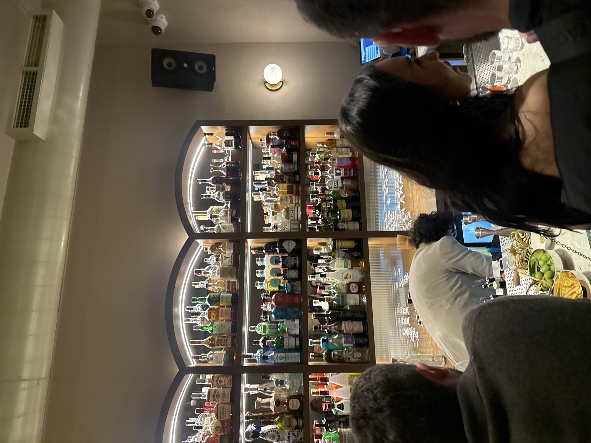 The image captures a bar setting, likely in Yerevan, Armenia. A wall-mounted speaker and ventilation shaft are visible on the left wall. A wall light is mounted above a large, arched liquor cabinet stocked with a variety of bottles, illuminated by integrated lighting. In the foreground, two individuals are partially visible, suggesting they are customers. A bartender, wearing a white shirt and dark gloves, is seen working behind the bar, which displays glassware and garnishes like limes. A television screen is visible behind the bartender.