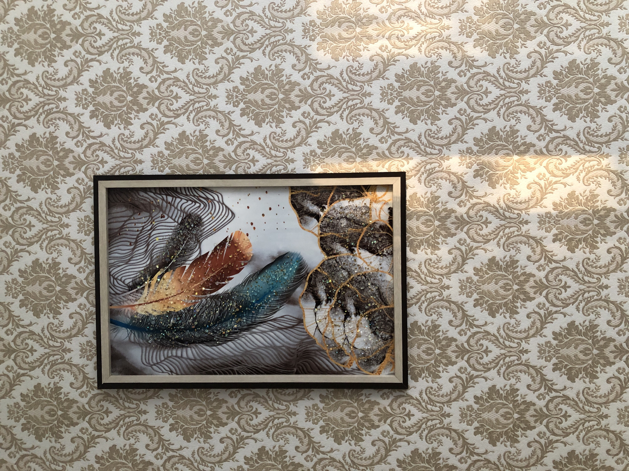 The image shows a framed piece of art hanging on a wall covered with damask wallpaper. The wallpaper has a repeating pattern of light beige floral and scroll motifs against a cream background. The framed art is rectangular with a dark frame and a lighter inner mat. The artwork itself depicts several feathers with iridescent details, appearing to be made of natural materials with added glitter or sequins. The feathers are in shades of brown, orange, and blue, and are layered against a background that includes abstract lines and textured elements of black, white, and gold. Sunlight casts a warm diagonal glow across the top right portion of the wallpaper and the artwork, suggesting it is daytime. The scene is static, with no activity or people visible.